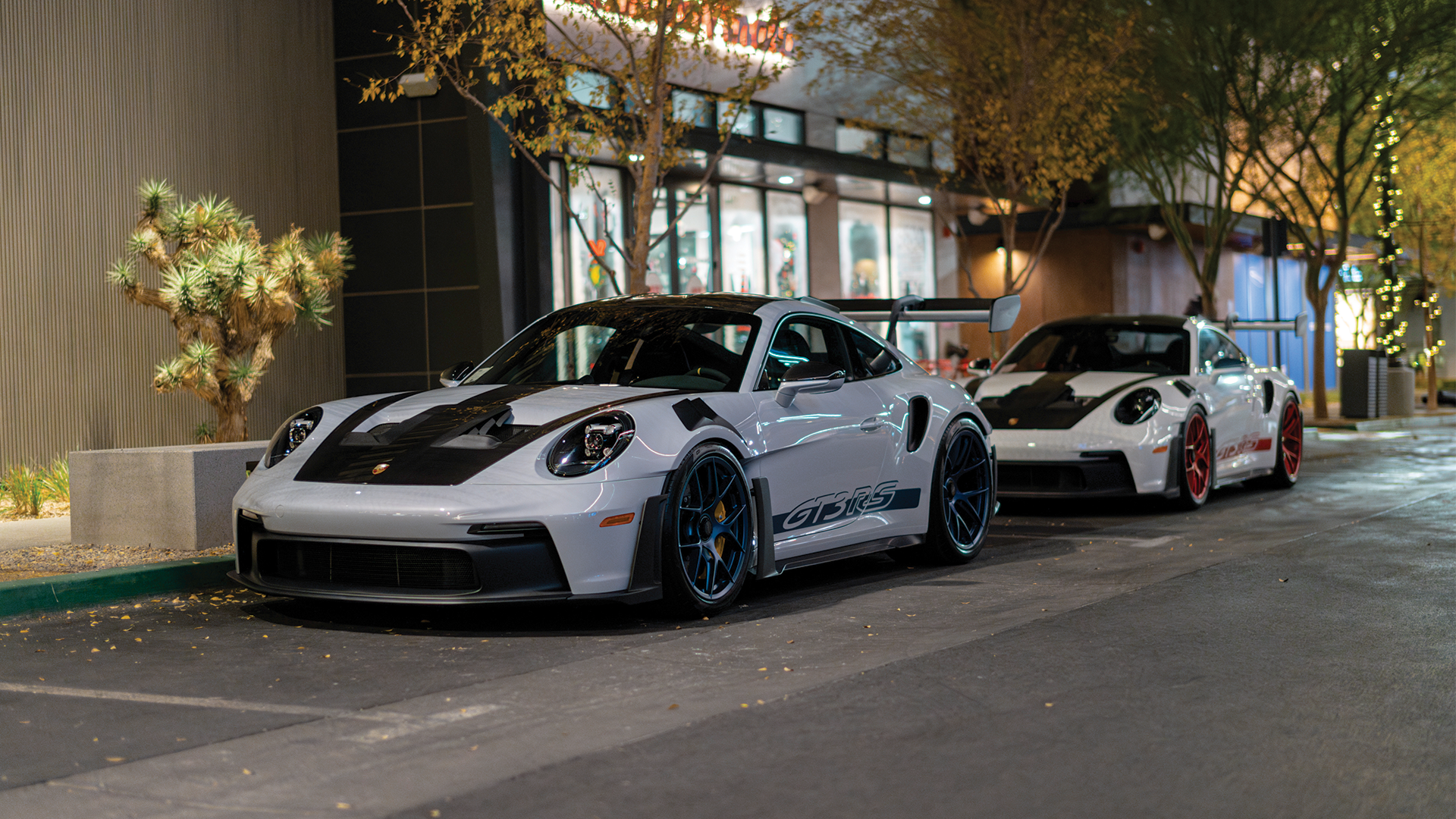 Two white sports cars parked on a city street at night, with trees decorated with lights and a storefront in the background.