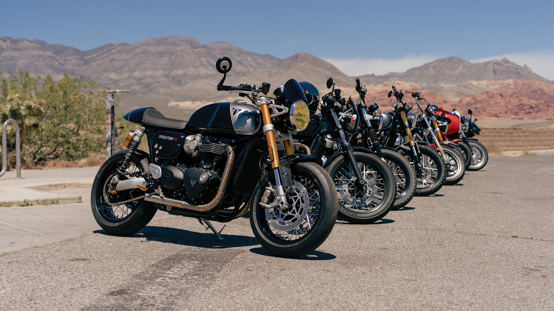 Multiple motorcycles parked in a row on a paved area with desert mountains in the background under a blue sky.