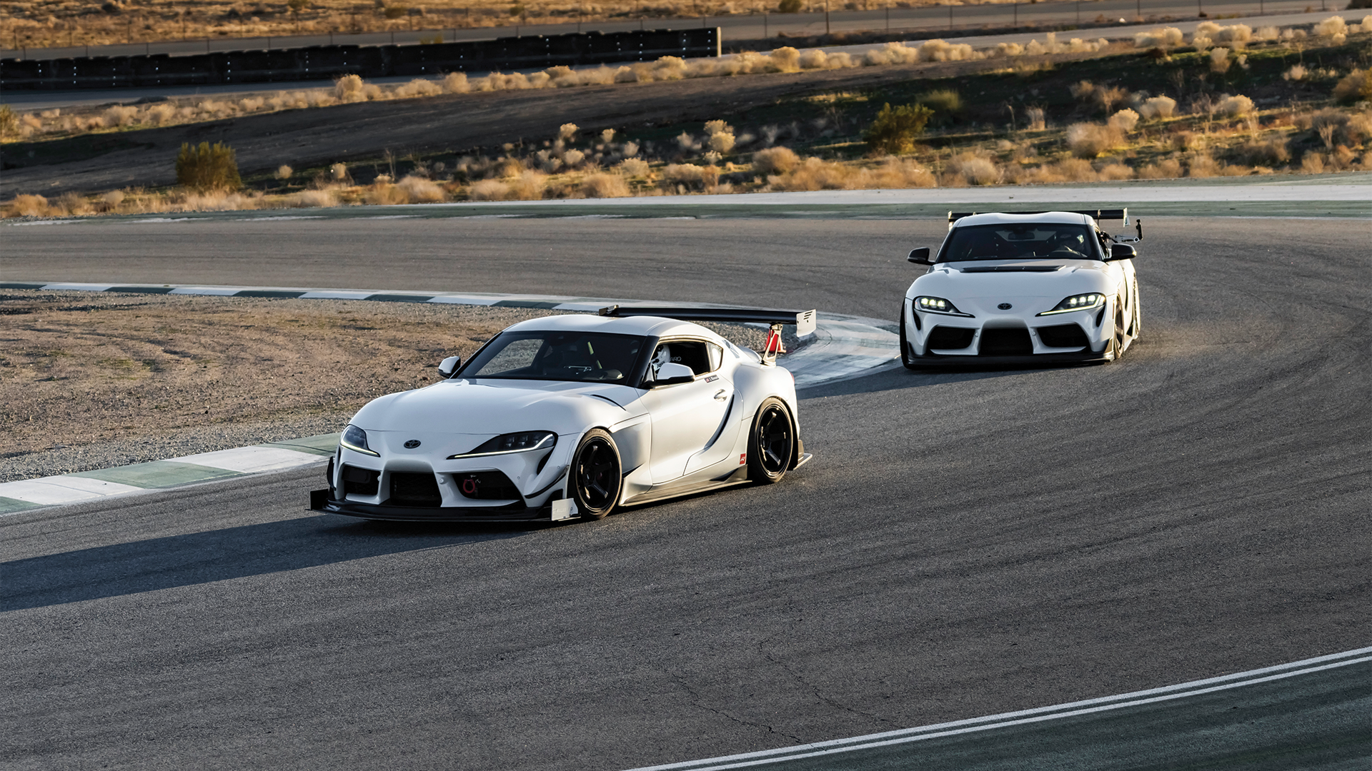 Two white sports cars on a race track, one in the foreground and one in the background, with a desert landscape and hills in the distance.