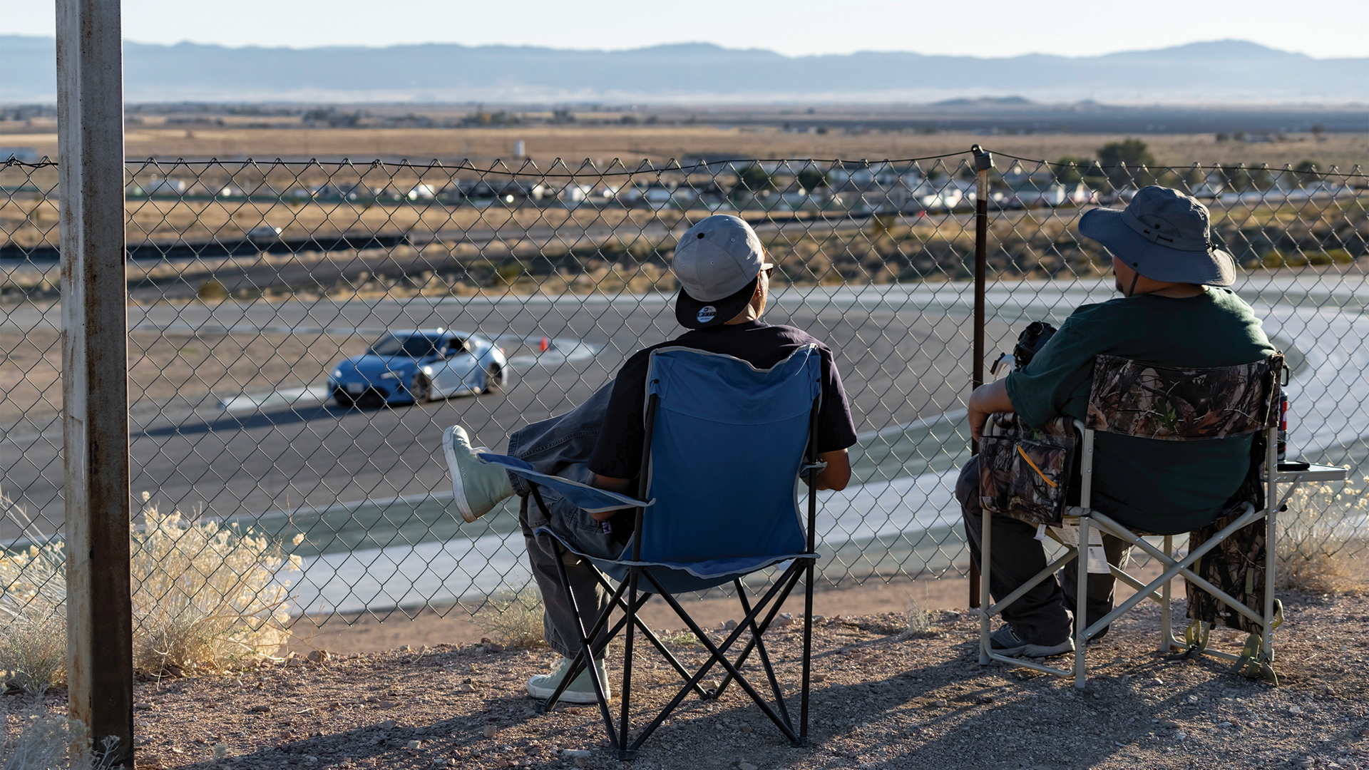 Two people sitting in folding chairs and watching a car race behind a chain-link fence, with a desert landscape and mountains in the background.