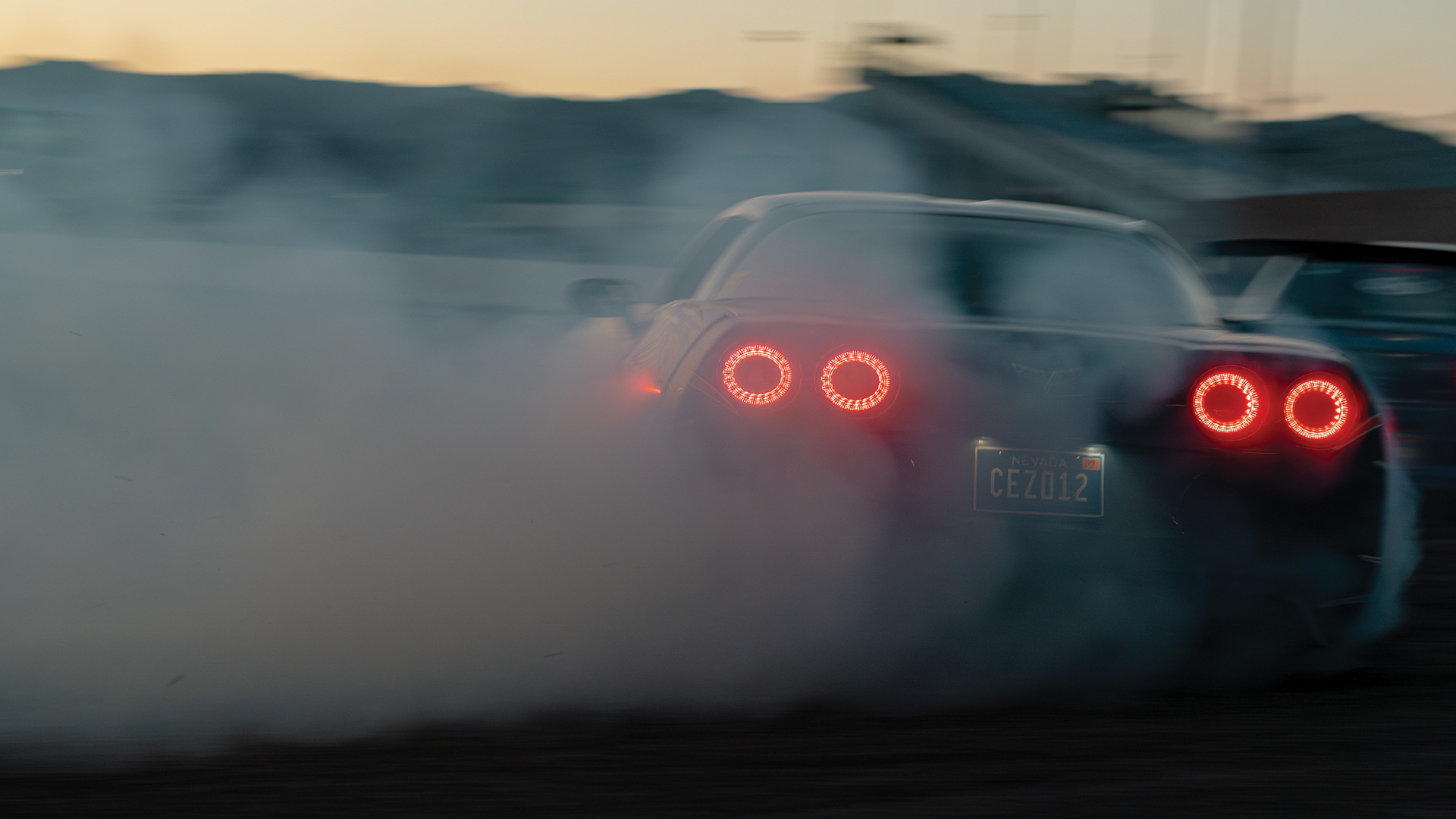 A sports car with Nevada license plate number CEZ012 is drifting, creating smoke from tires, in a possibly controlled stunt or race setting during dusk.