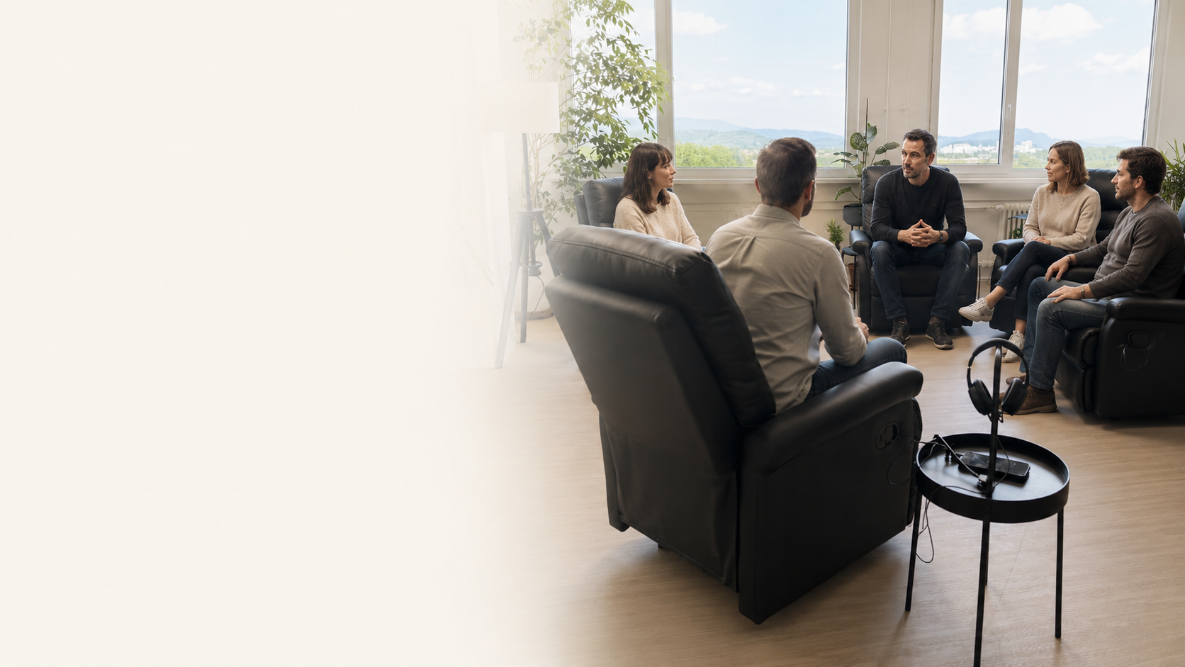 Group of six people sitting in a semi-circle conversation in a bright office with large windows, indoor plants, and a view of mountains.