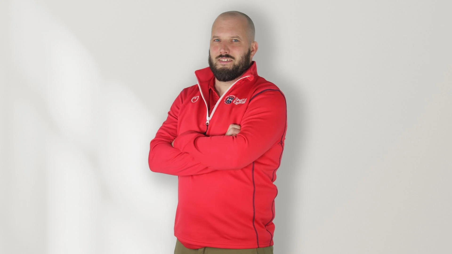 A man with a beard and shaved head wearing a red sporty jacket with a zipper, standing with arms crossed against a plain light-colored wall.