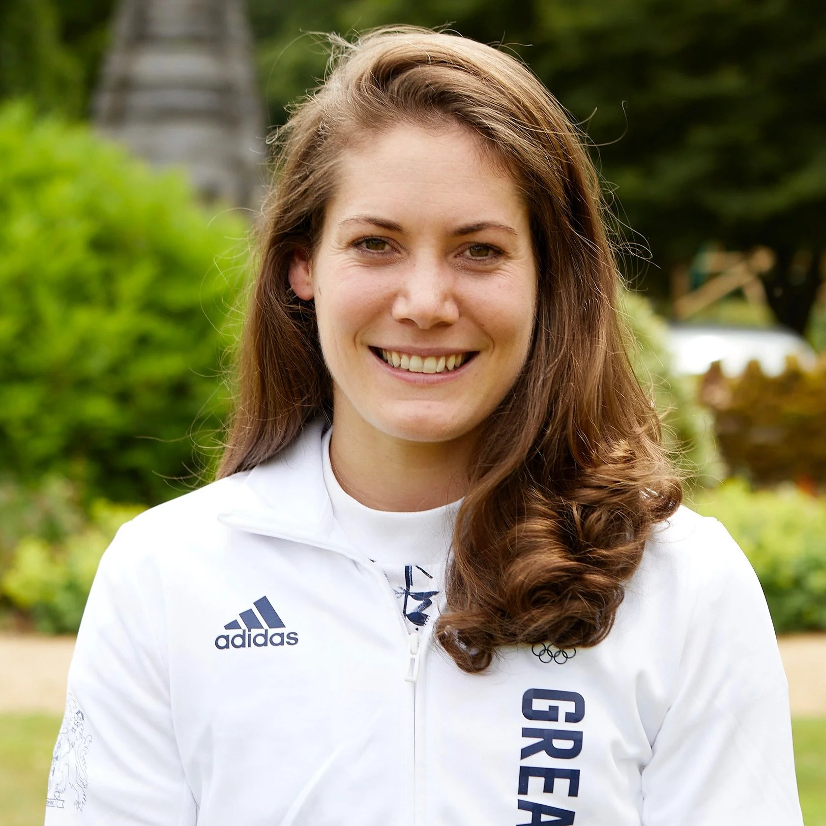 A smiling woman with long, curly brown hair outdoors, wearing a white athletic jacket with the Adidas logo and the word 'Great Britain' on it.