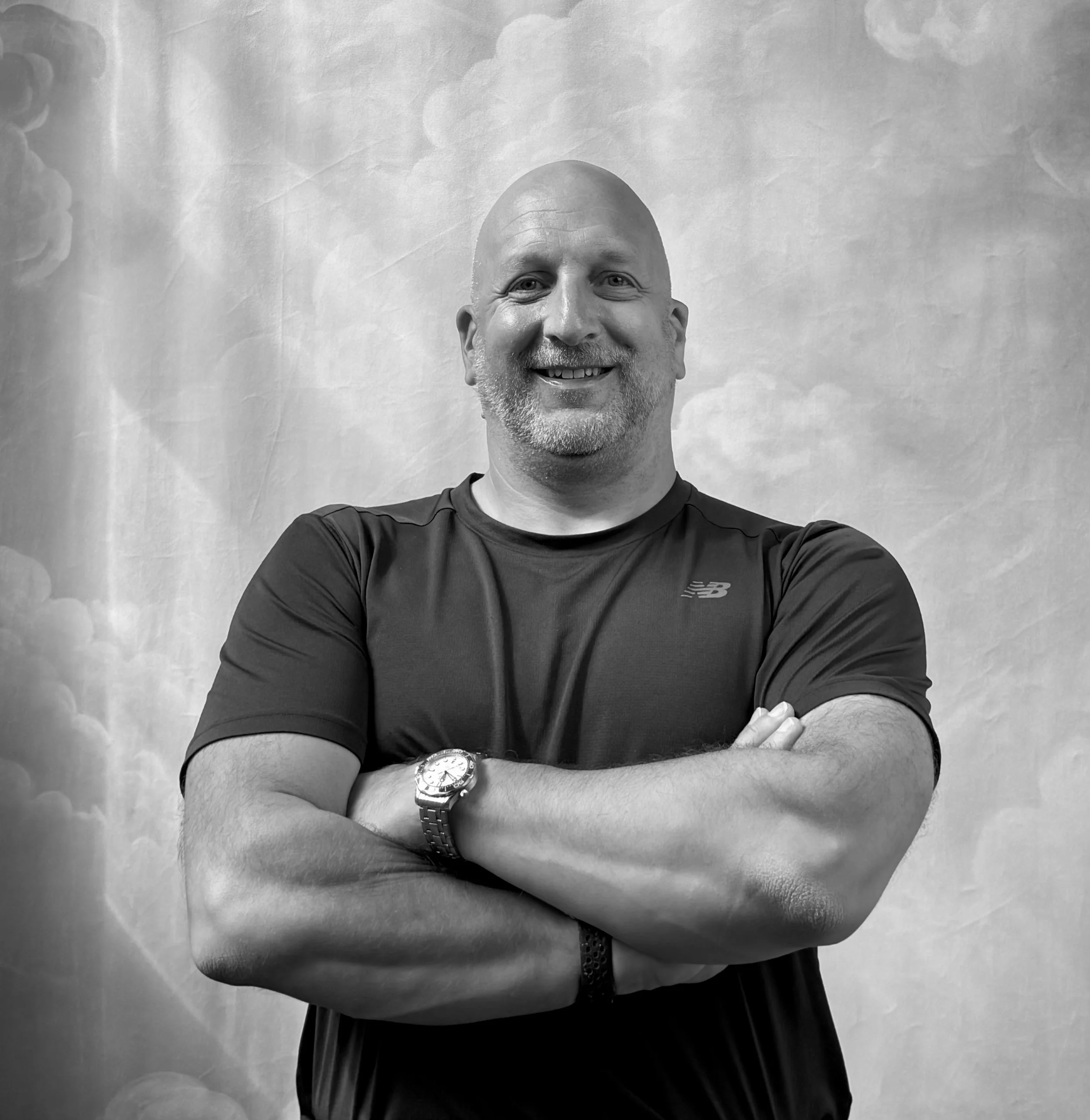Black and white photo of Mark Skimming a smiling, bald man with a beard,  standing in front of a cloudy sky background with clouds and some blurred circular shapes, with his arms crossed.