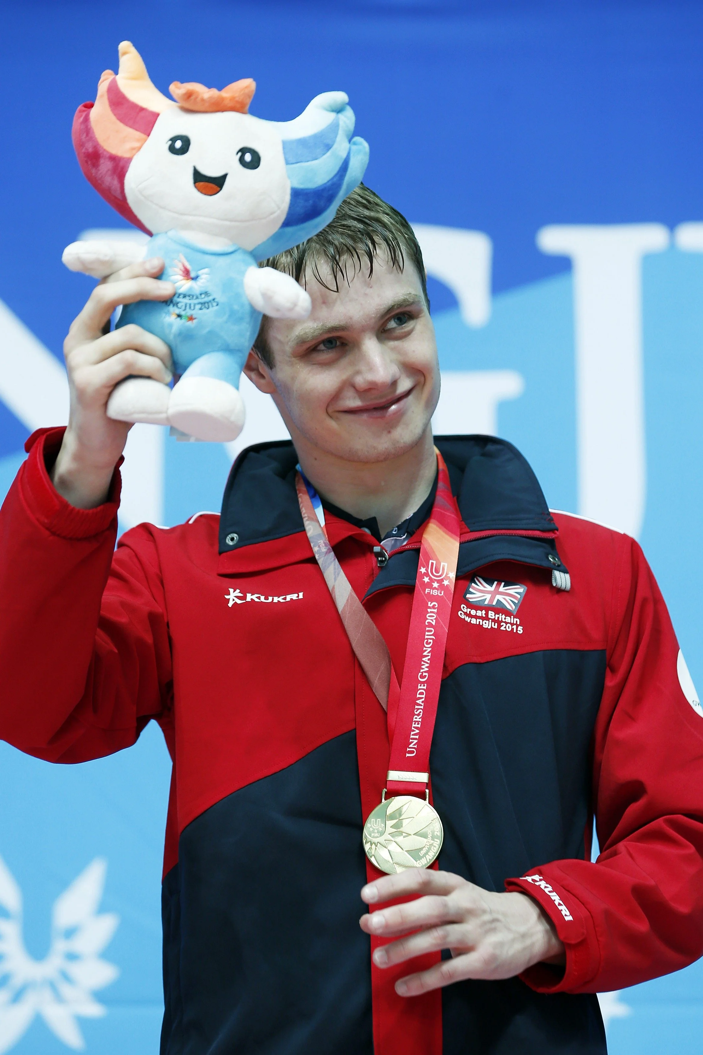 A male athlete in a red jacket with medals around his neck, holding a plush mascot of the Gwangju 2015 Summer Universiade, smiling after winning a medal.