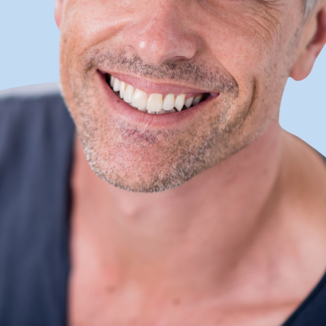 Close-up of a smiling man with facial stubble, wearing a dark shirt against a light blue background.