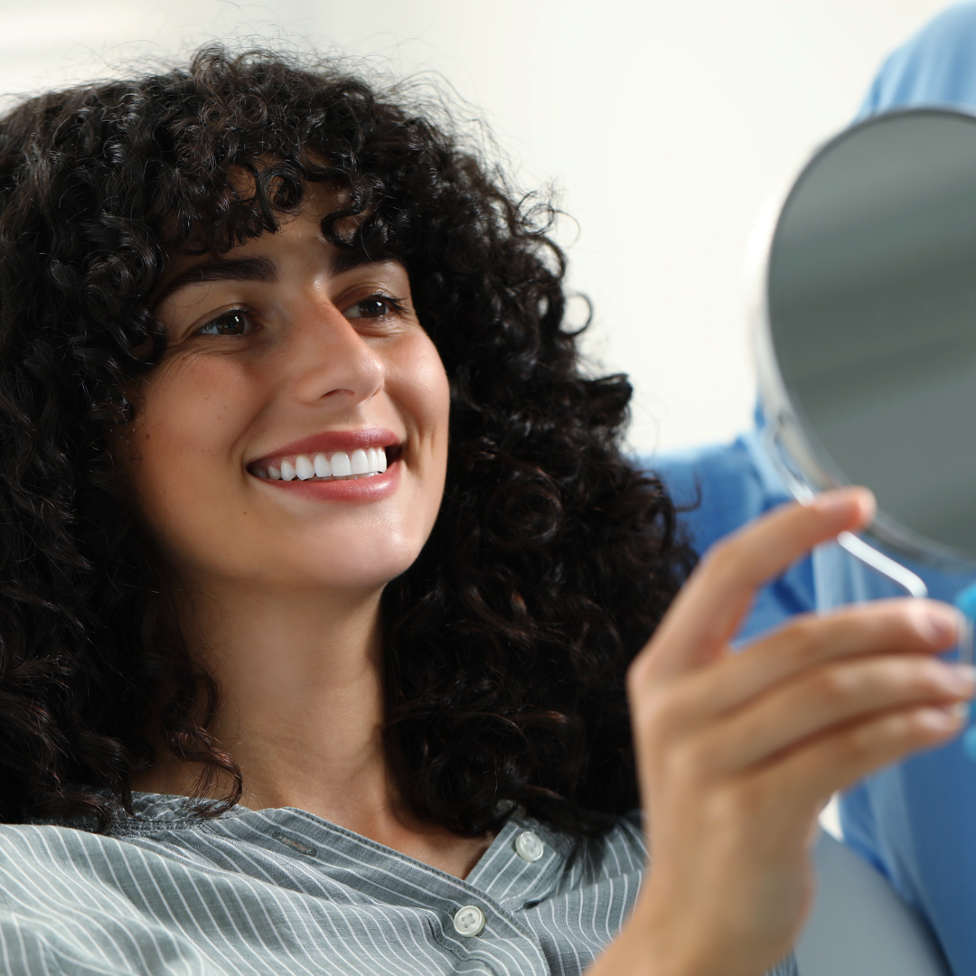 A woman with curly hair smiling and holding a mirror in a dental setting.