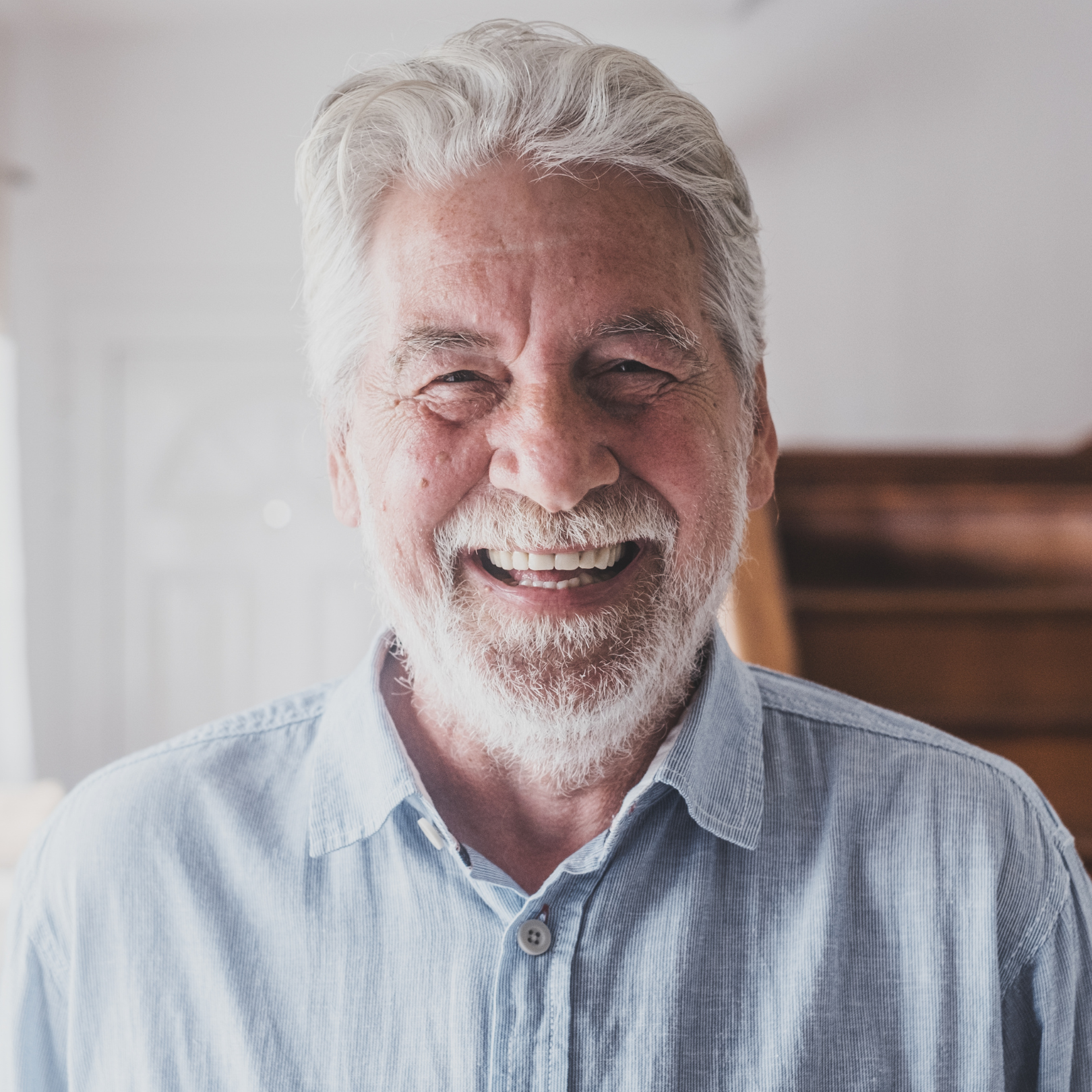 Smiling elderly man with white hair and beard wearing a light blue shirt indoors.