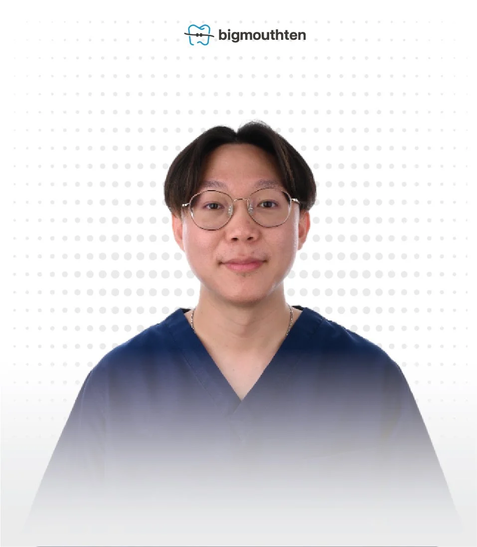 A young man with glasses and dark hair, wearing a dark blue medical scrub, smiling at the camera, standing against a white background with dotted pattern and the 'bigmouthten' logo at the top.