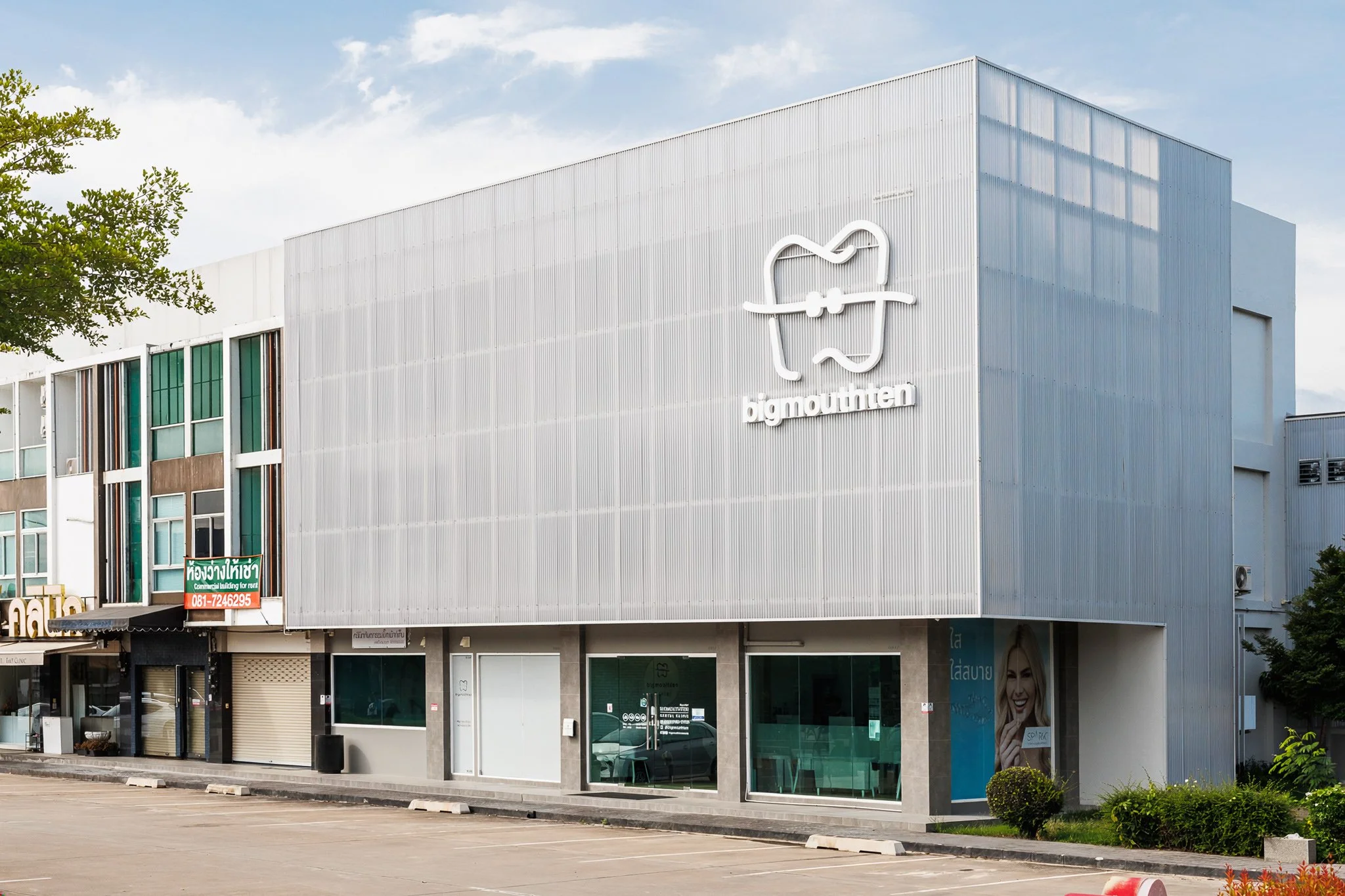 Modern commercial building with a logo of a smiley face in a shopping bag, and the word 'bigmauthen' beneath it. The building has a sleek exterior with large glass windows and an empty parking lot in front.