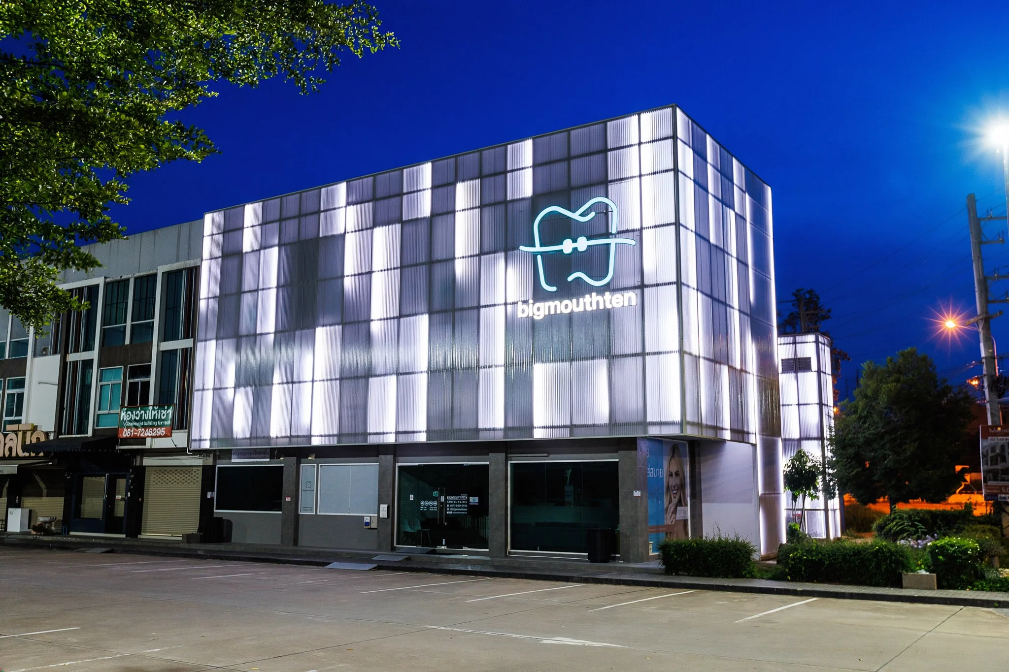 A modern building with a large illuminated sign featuring a stylized tooth and the text 'bigmouthten' in lowercase. The building's facade is made of glowing panels, and the scene appears to be at dusk with a clear sky. There are trees and streetlight