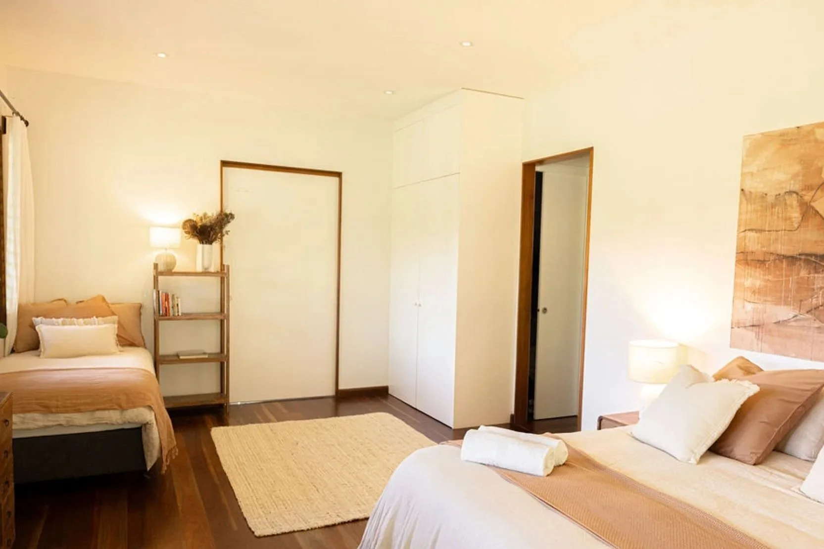 A bedroom with two beds, one on the left and one on the right, lamps, and a wooden floor. There is a doorway and a closet, with a small table and a shelf with books and a vase with flowers.