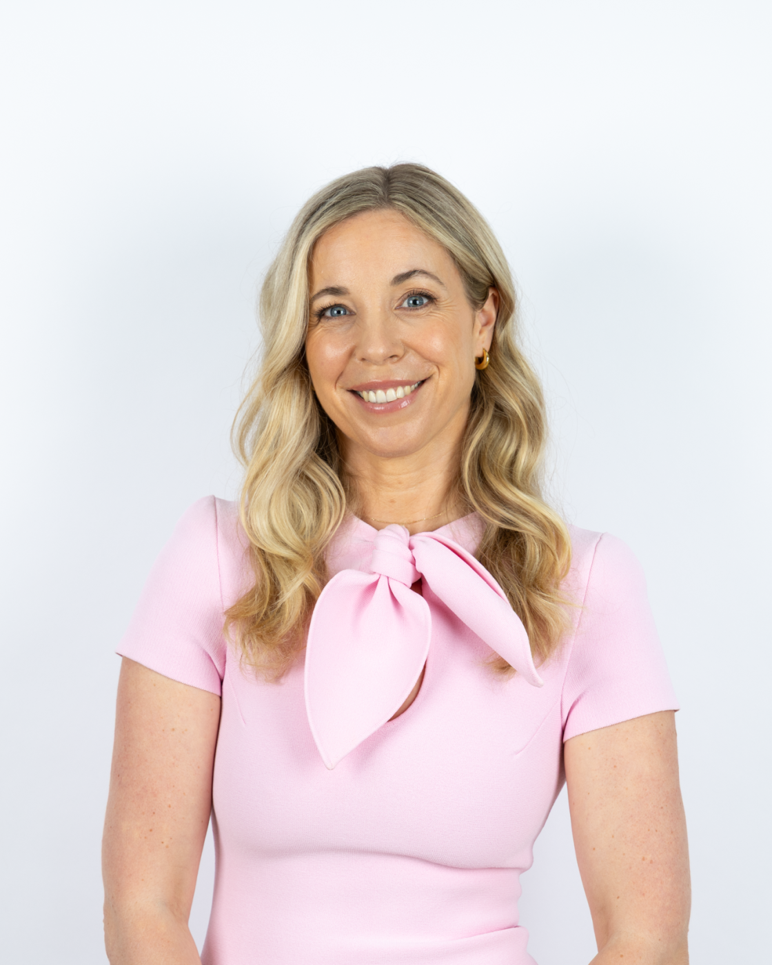 Smiling woman with blonde hair wearing a light pink dress with a bow tie collar, against a white background.