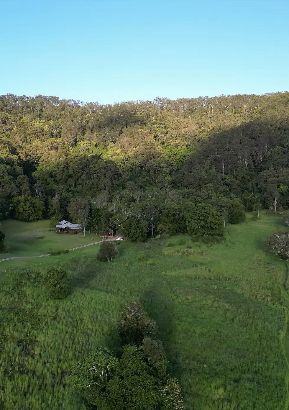 A house near a wooded hillside with a bright blue sky and green fields in the foreground.