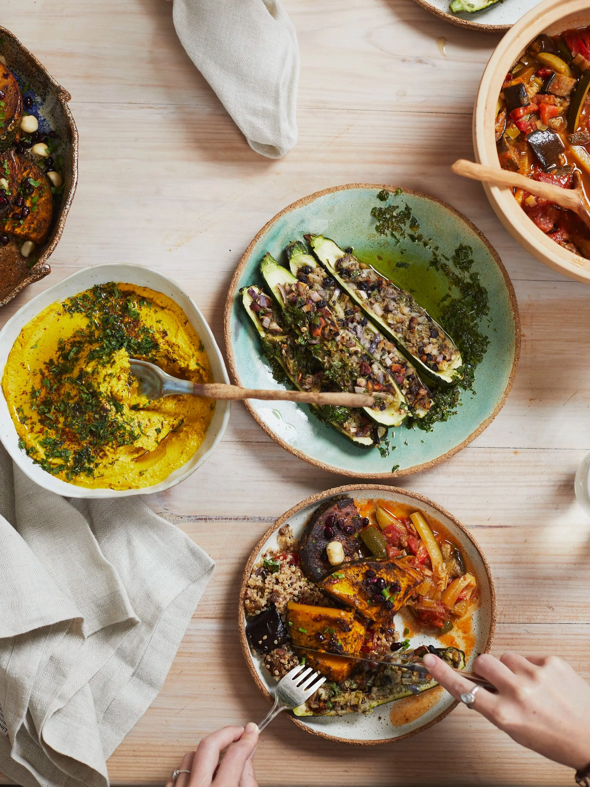 Various colorful dishes of roasted vegetables, hummus, and stuffed zucchini boats on a light wooden table, with a person's hands cutting into the stuffed vegetable with a fork and knife.