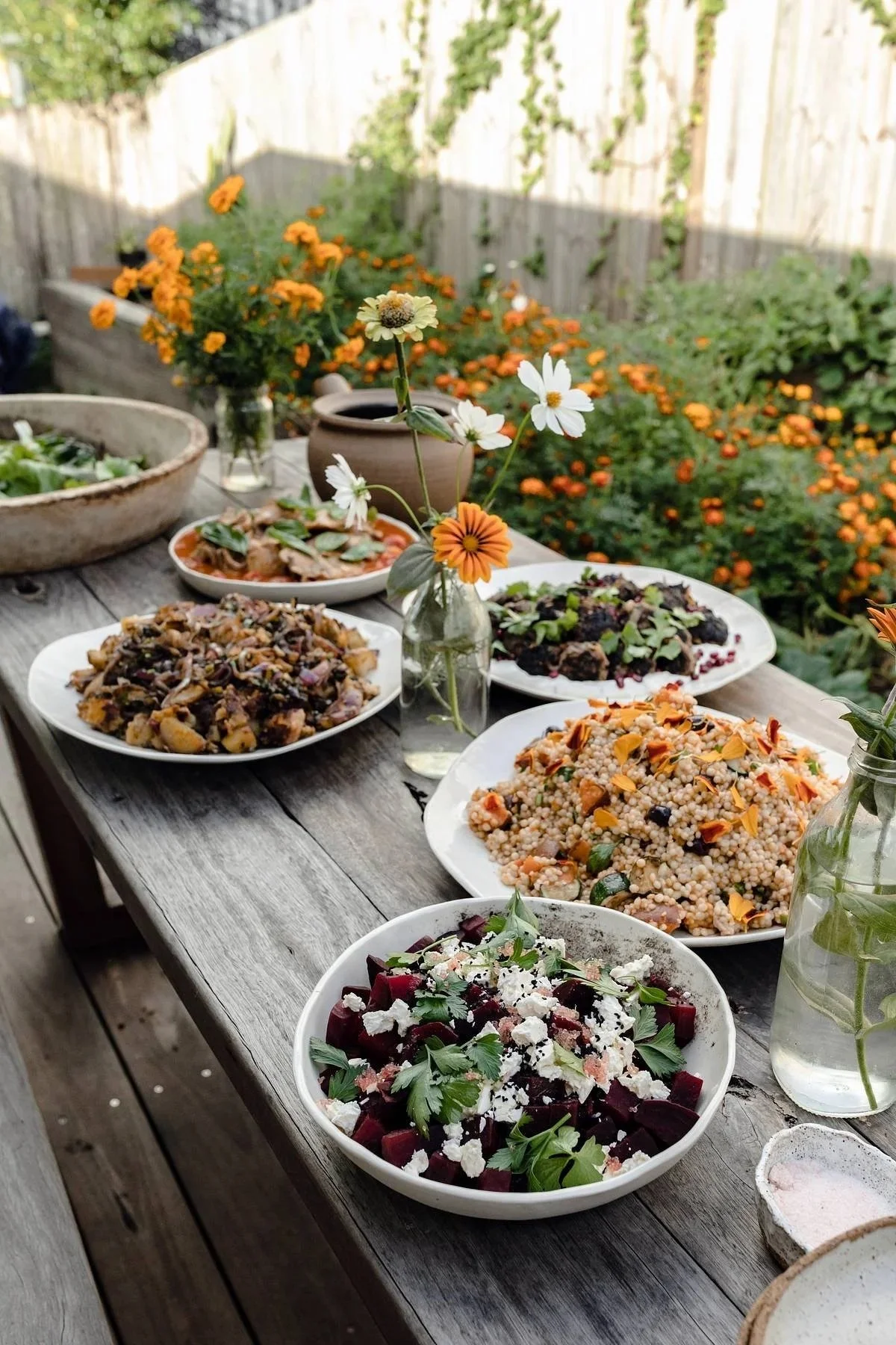 Outdoor wooden table with various dishes and small vases with flowers, set in a garden with orange flowers and a wooden fence in the background.
