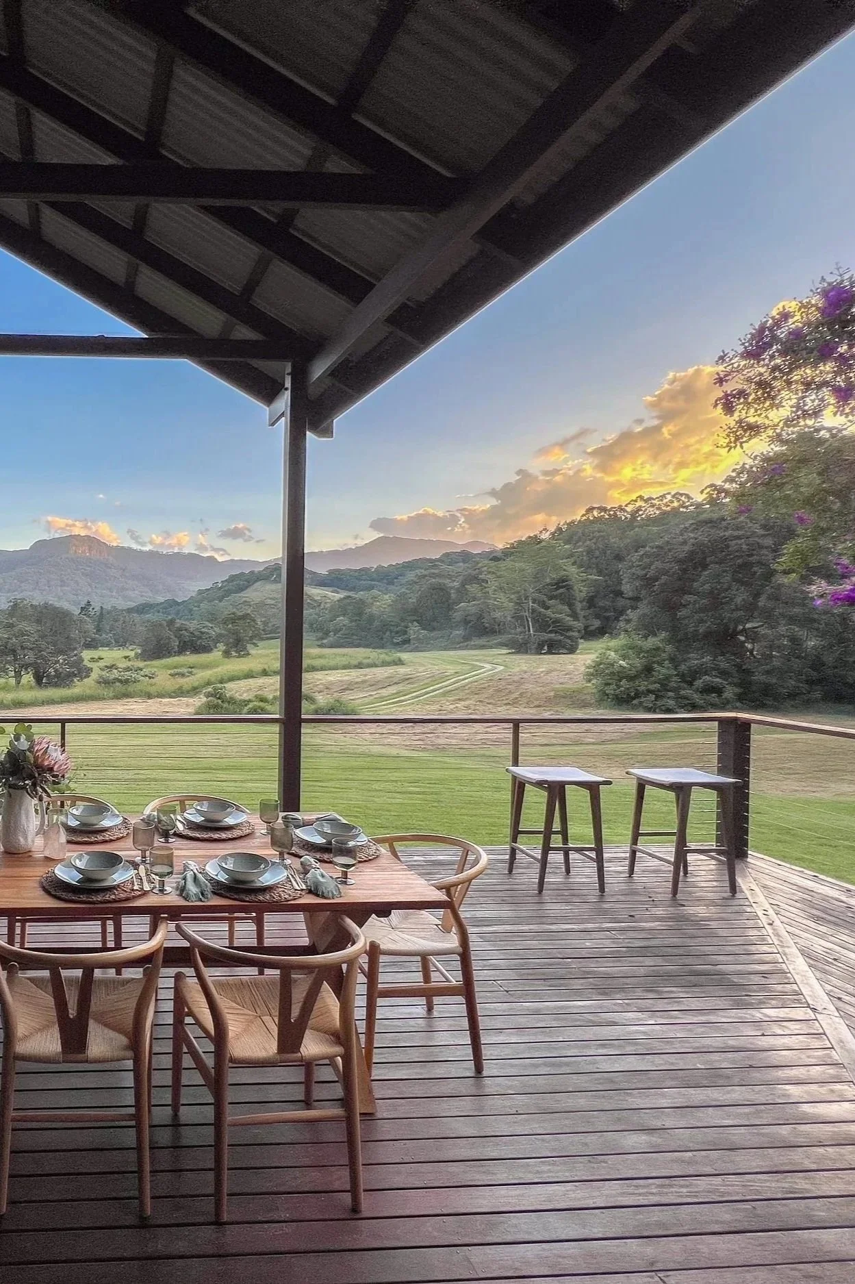 A balcony overlooking a scenic landscape with green hills, trees, and mountains at sunset. The balcony has a wooden dining table set for a meal, with chairs, placemats, dishes, and glasses. Two stools are positioned near the railing, and the roof of the balcony extends overhead.