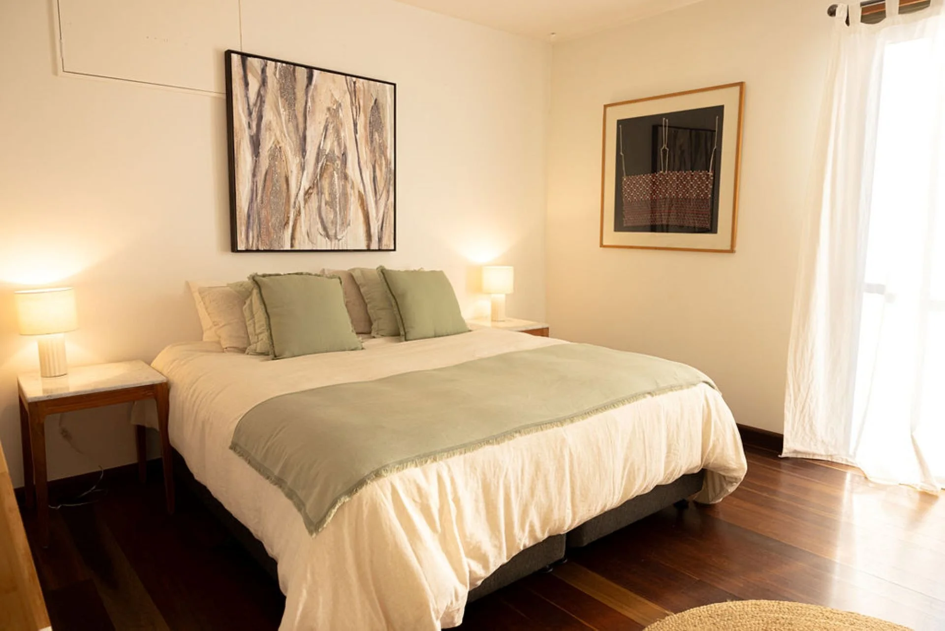 A cozy bedroom with a bed covered in a light cream quilt with green accents, two matching bedside lamps, and artwork hanging on the cream-colored walls near a window with curtains.