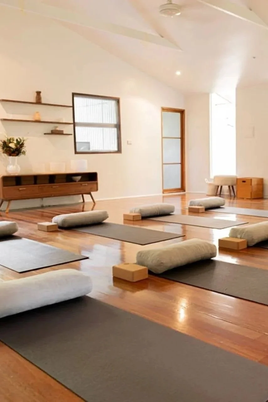 Yoga mats with bolsters and blocks arranged in a spacious, well-lit room with wooden floors, shelves, and minimal decor.