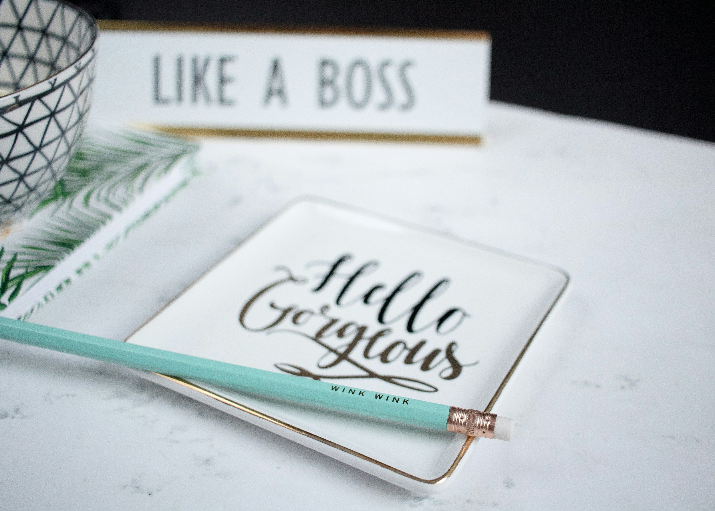A marble tabletop with a notebook that says "Hello Gorgeous," a teal pencil labeled "Wink Wink," a patterned bowl, a card with "Like a Boss," and some tropical patterned paper.