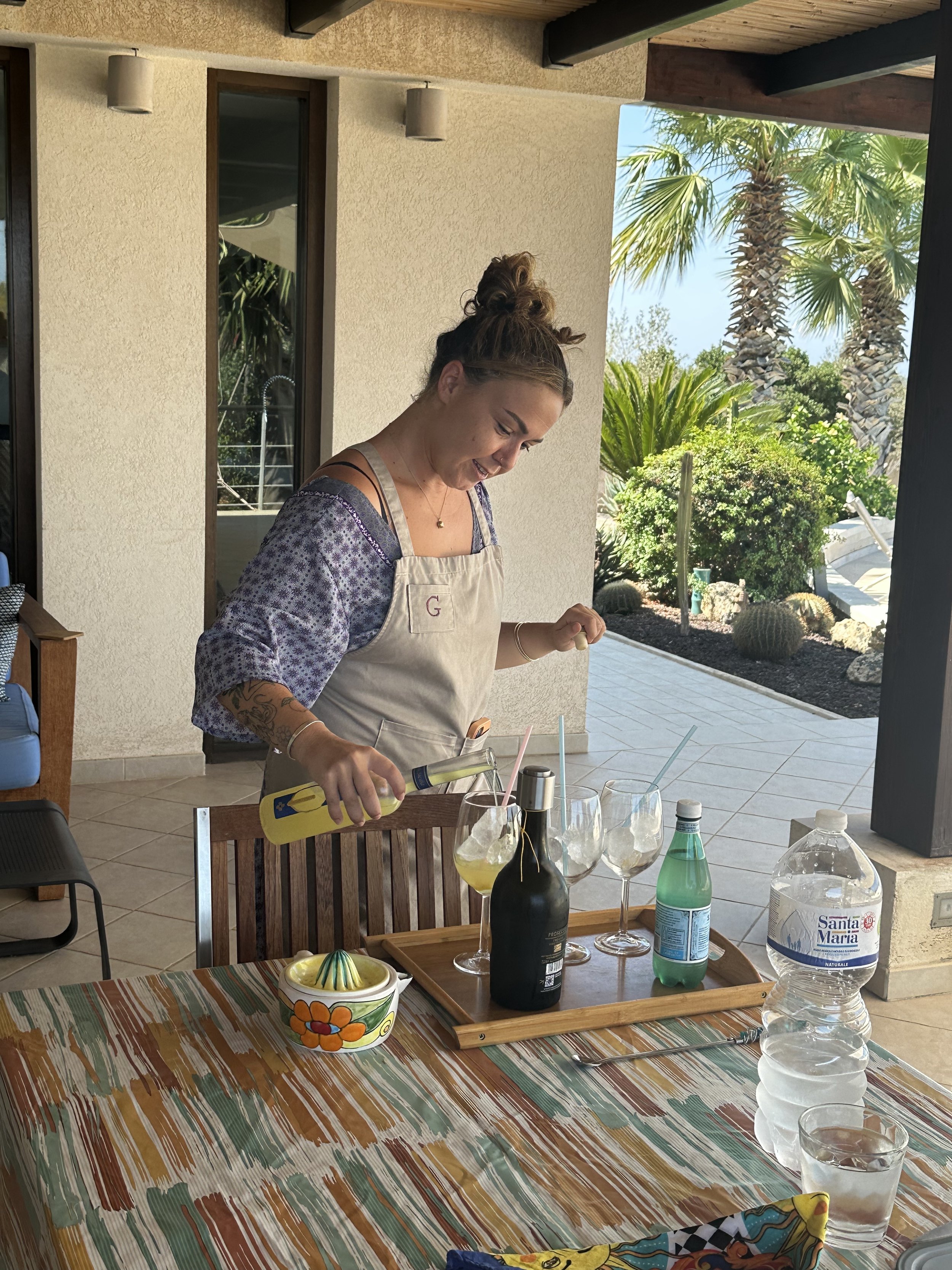 A woman in an apron pouring drinks into glasses on a table. The setting appears to be an outdoor patio with tropical plants in the background. On the table are bottled beverages, a citrus juicer, and glassware.