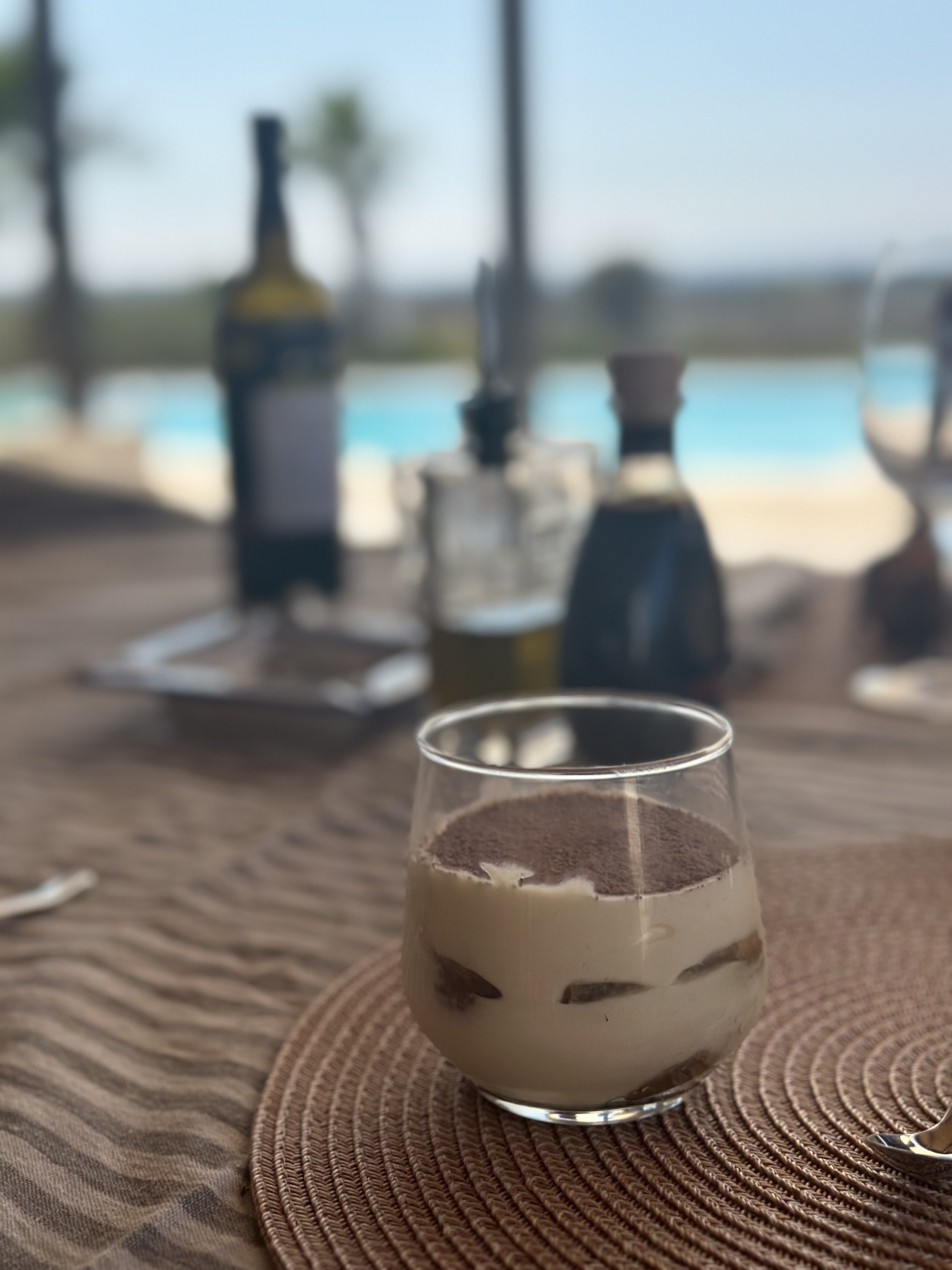 A dessert, likely tiramisu, in a glass on a woven placemat with blurred background of a table setting, bottles, and a pool.