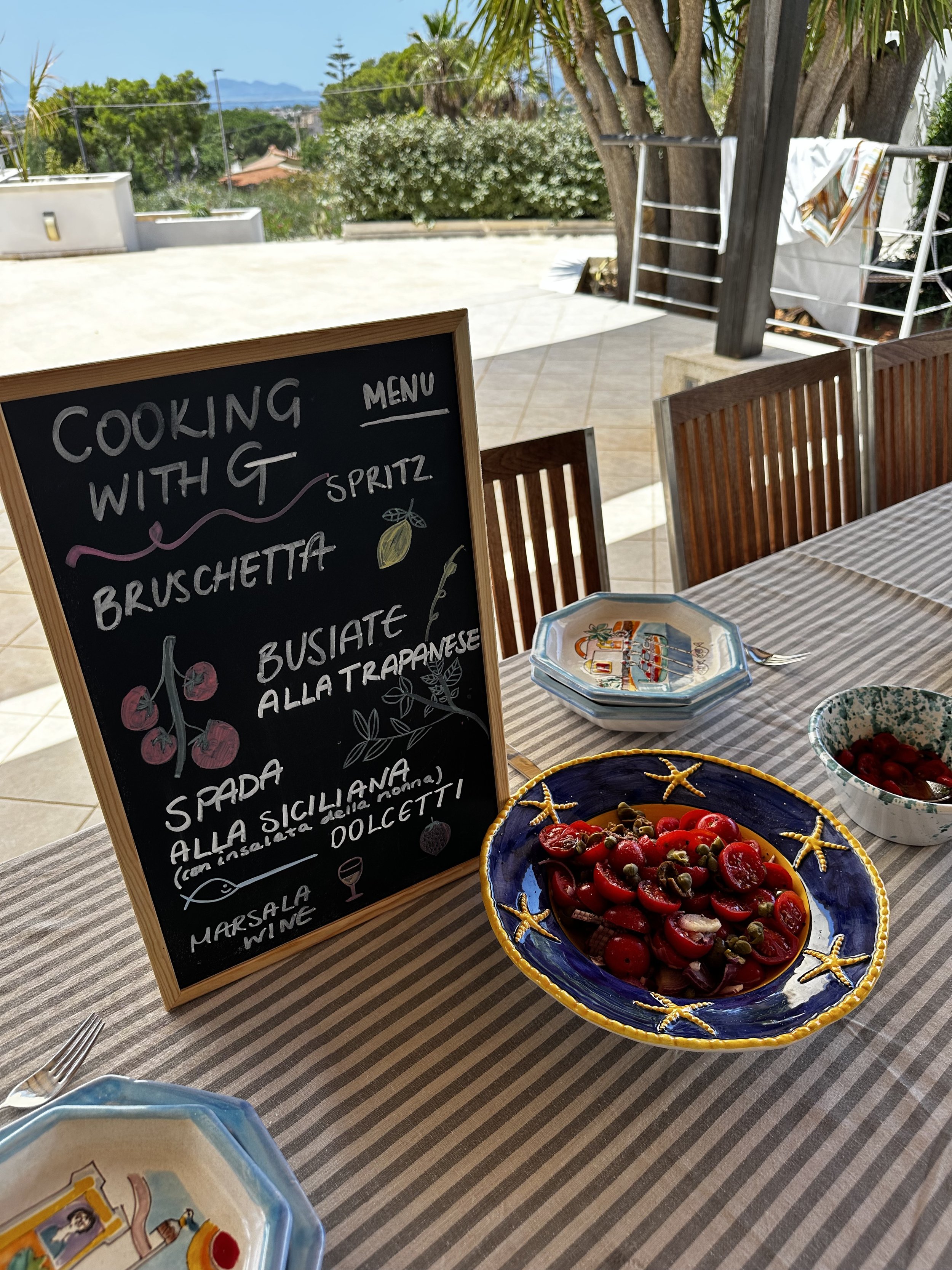 Outdoor table with striped cloth, decorative dishes, salad bowl, and chalkboard menu with Italian dishes.