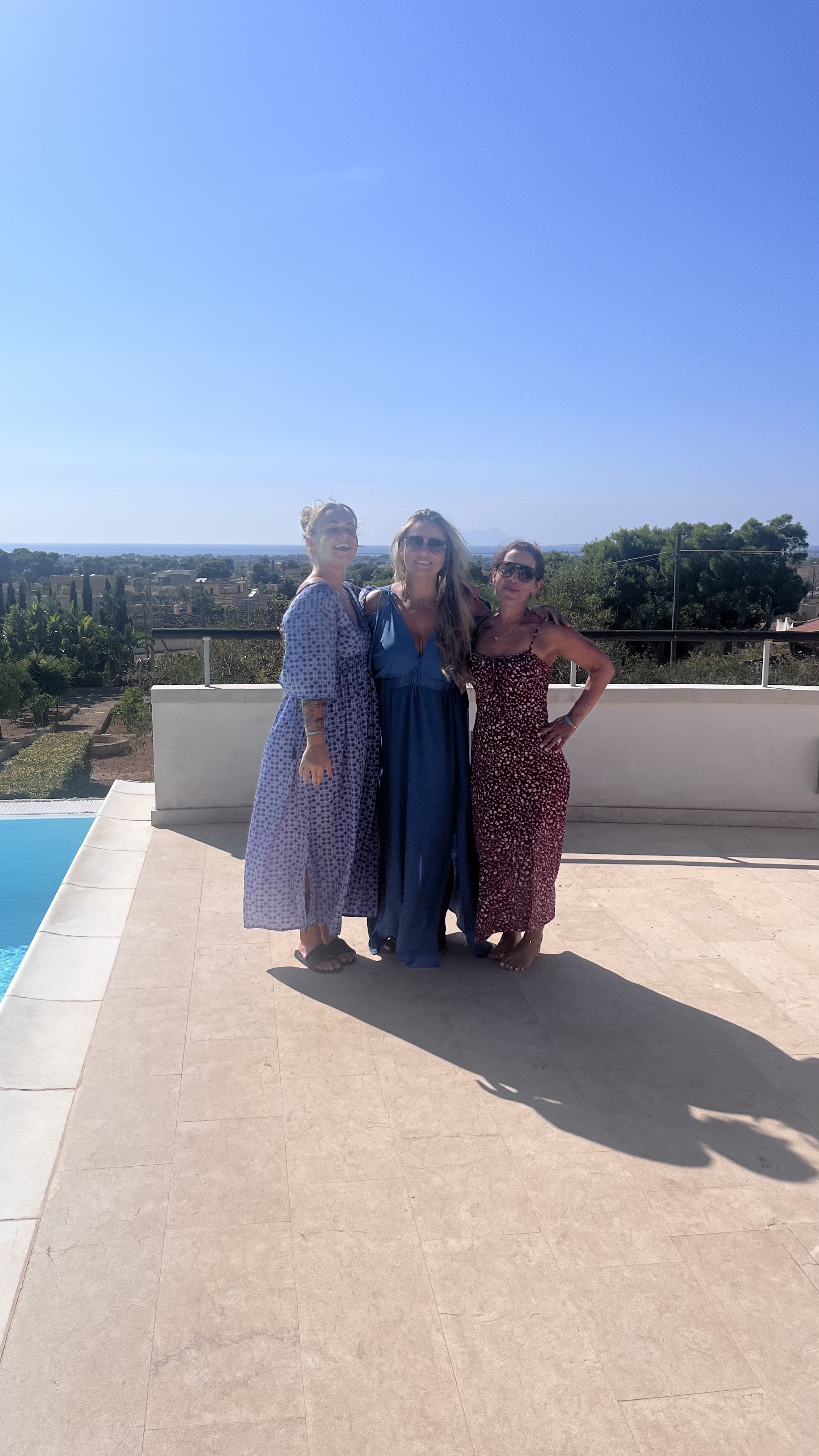 Three people in dresses standing on a terrace near a pool with a scenic view in the background.
