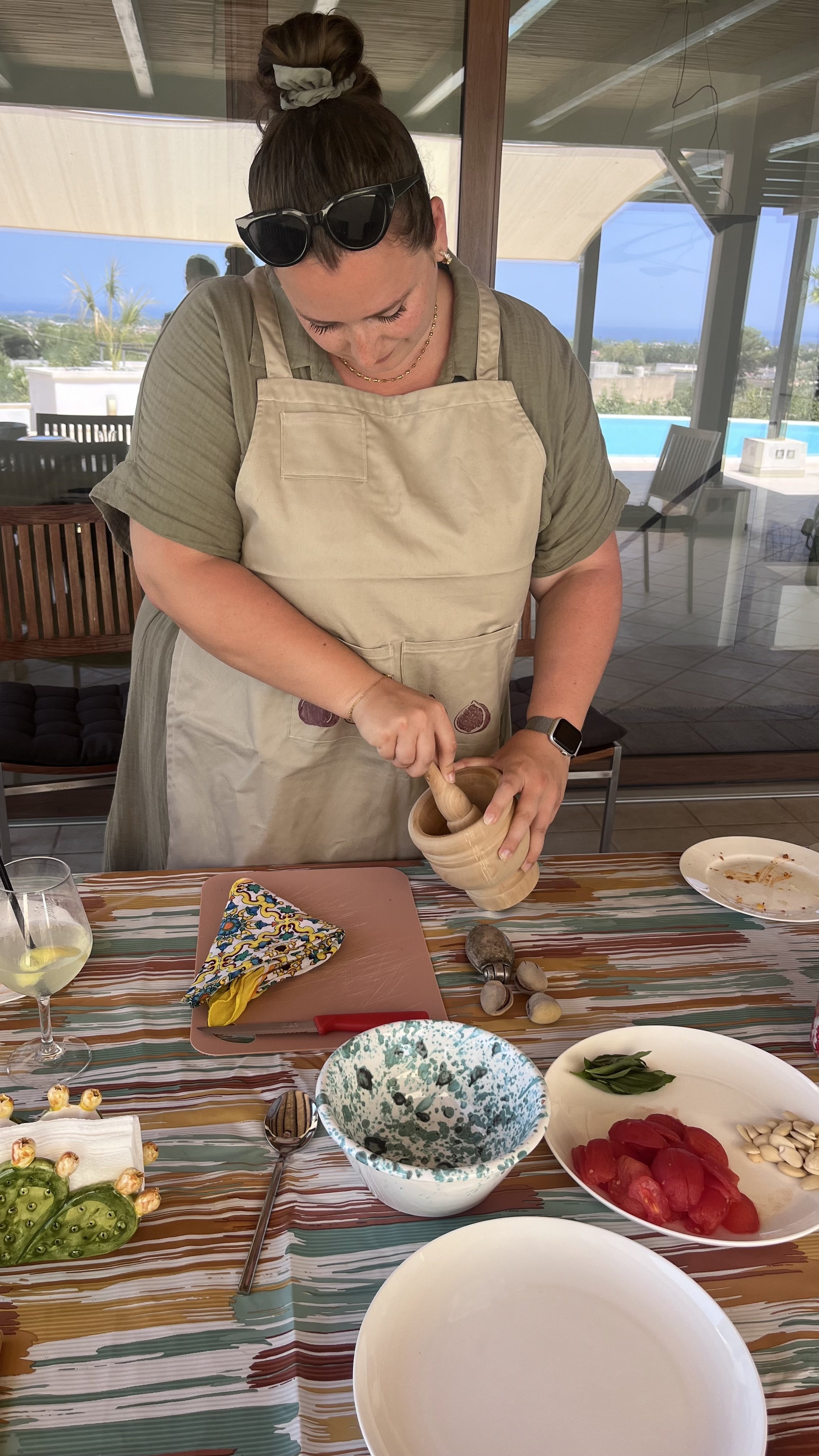 Person using a mortar and pestle on a table with kitchen items and ingredients, including tomatoes, basil, and garlic.