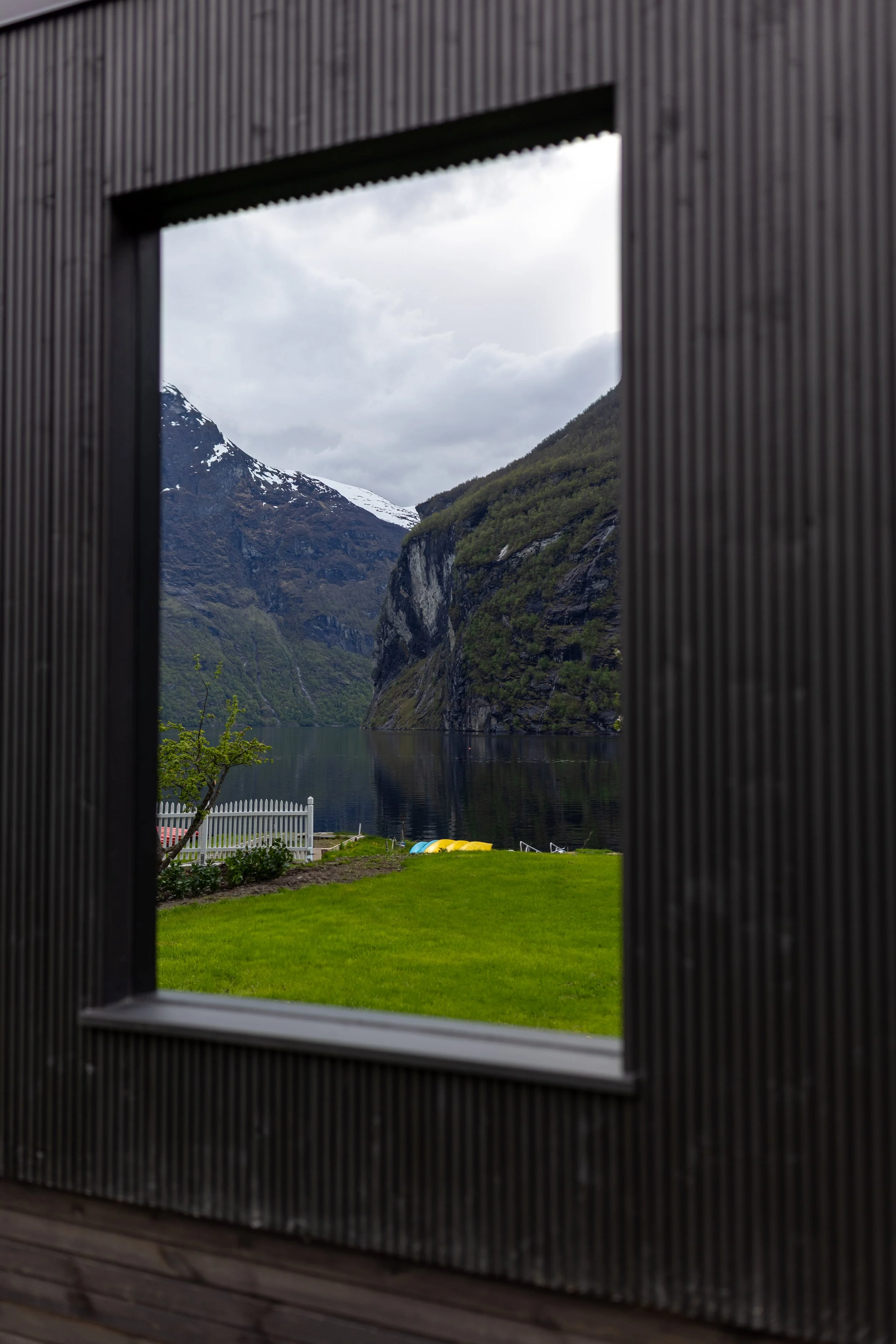 Utsikt gjennom et vindu av fjell, innsjø, grønn hage, tre, hvit hekk og fjell med snø i bakgrunnen.