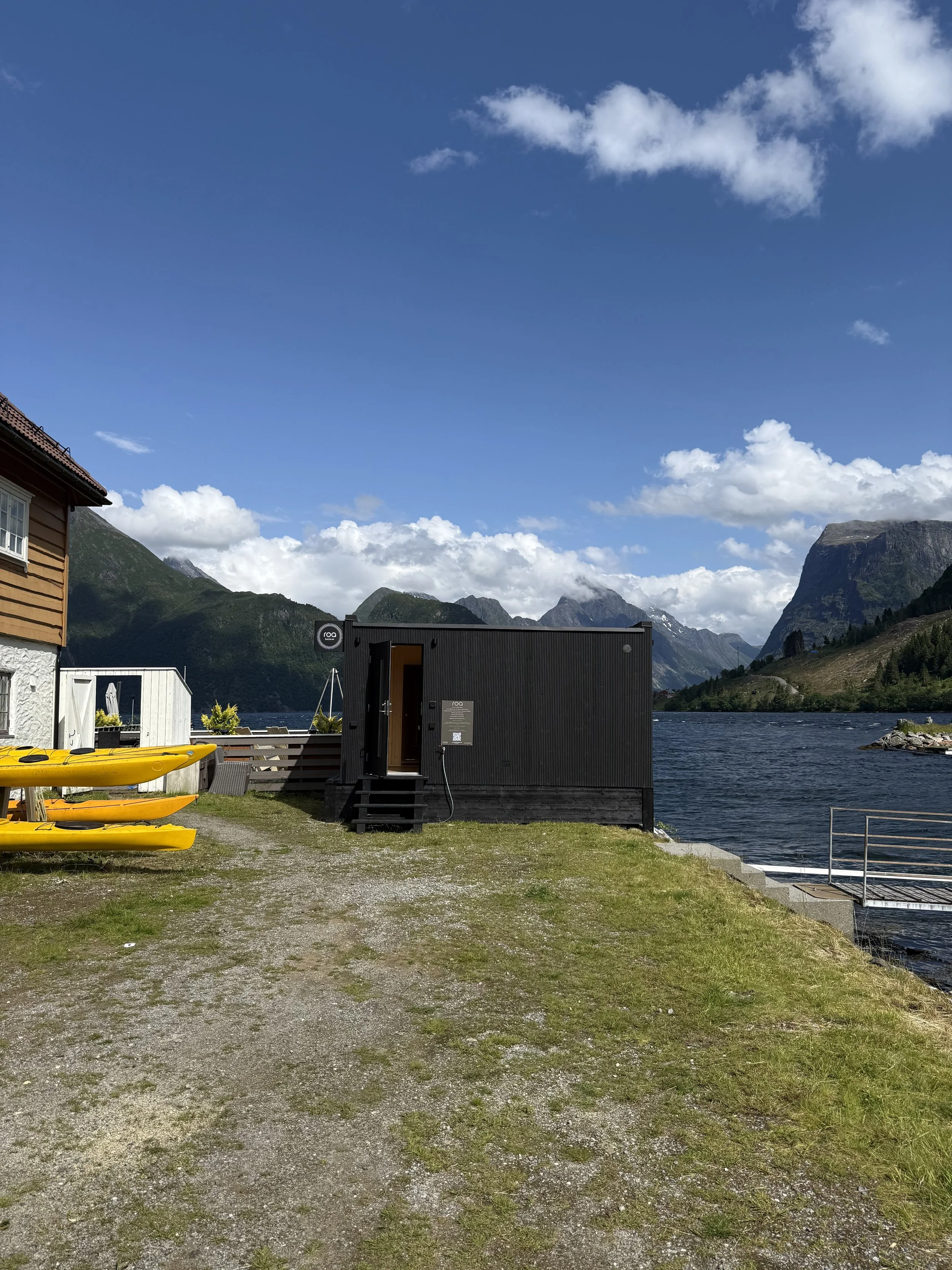 Badehytte ved fjorden med kajakker og fjell i bakgrunnen, sol og skyer på himmelen.