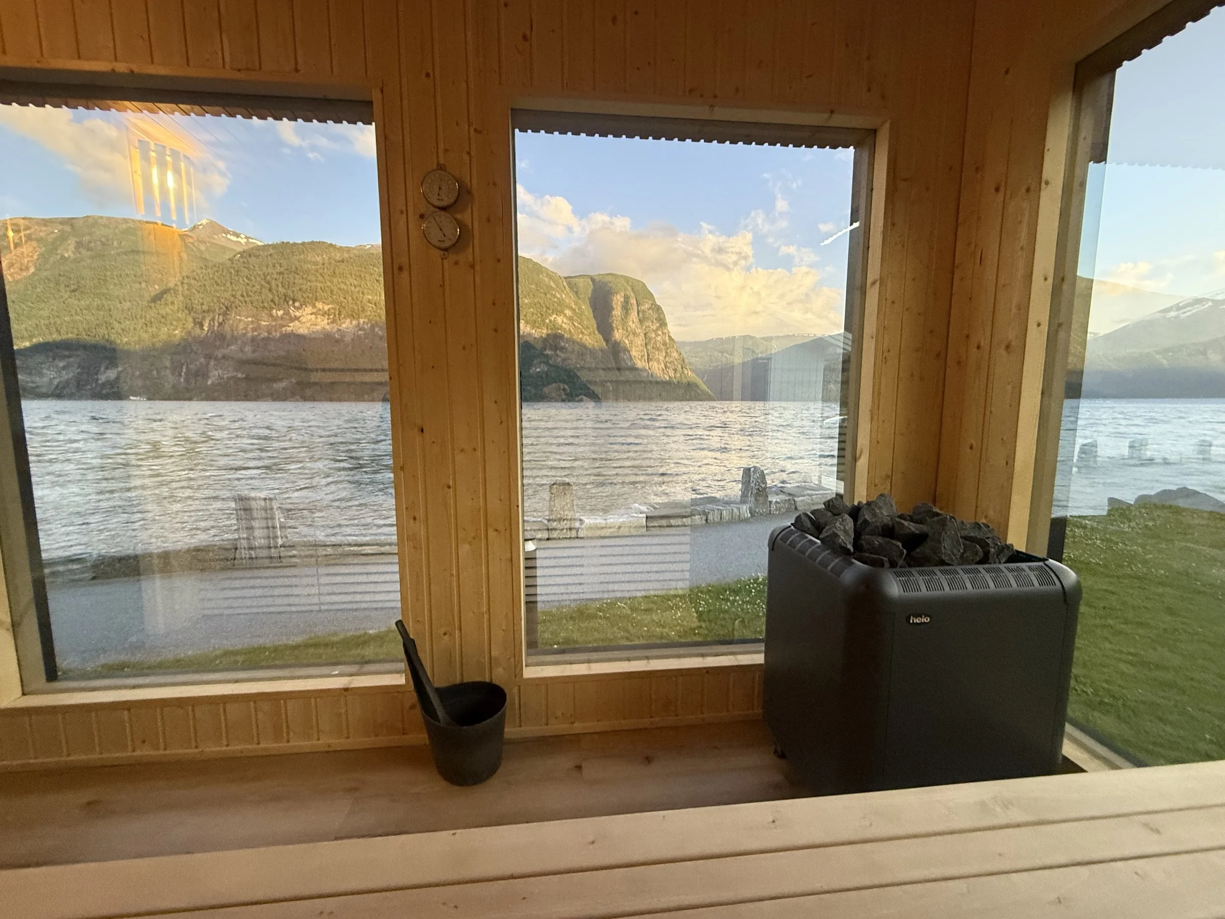 Innendørs med utsikt over en innsjø og fjell, vinduer, en liten svart ovn med stein på toppen, og en svart kopp.