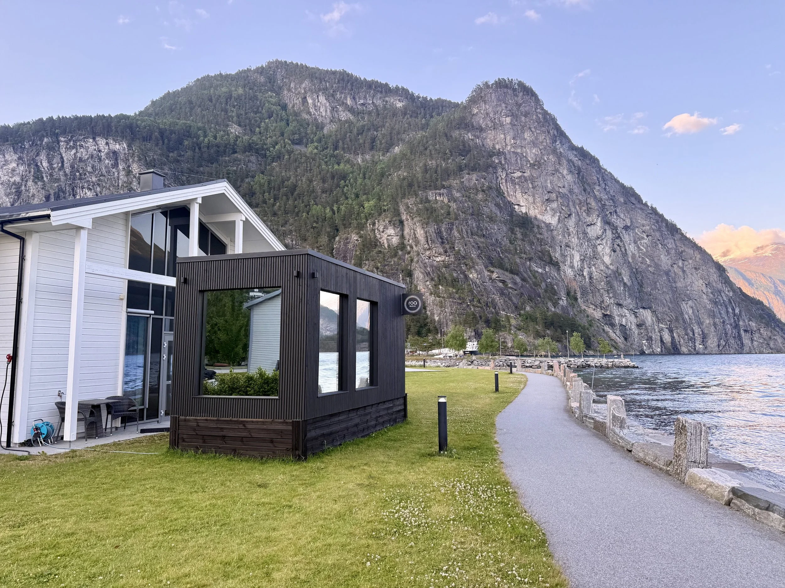 En bolig ved vannkanten med fjell i bakgrunnen, en gruslagt promenade og grønt gressområde.
