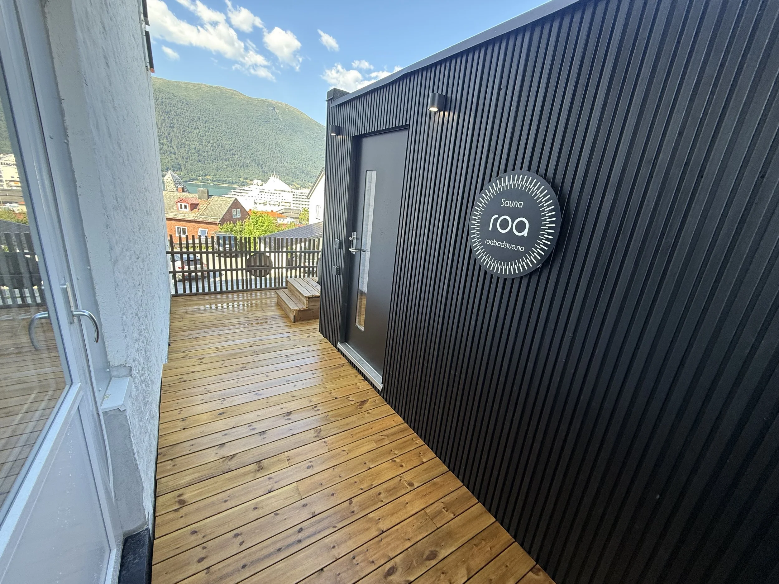 Utendørs ved sauna med svart vegg, treterrasse, og utsikt over fjell og by i bakgrunnen.