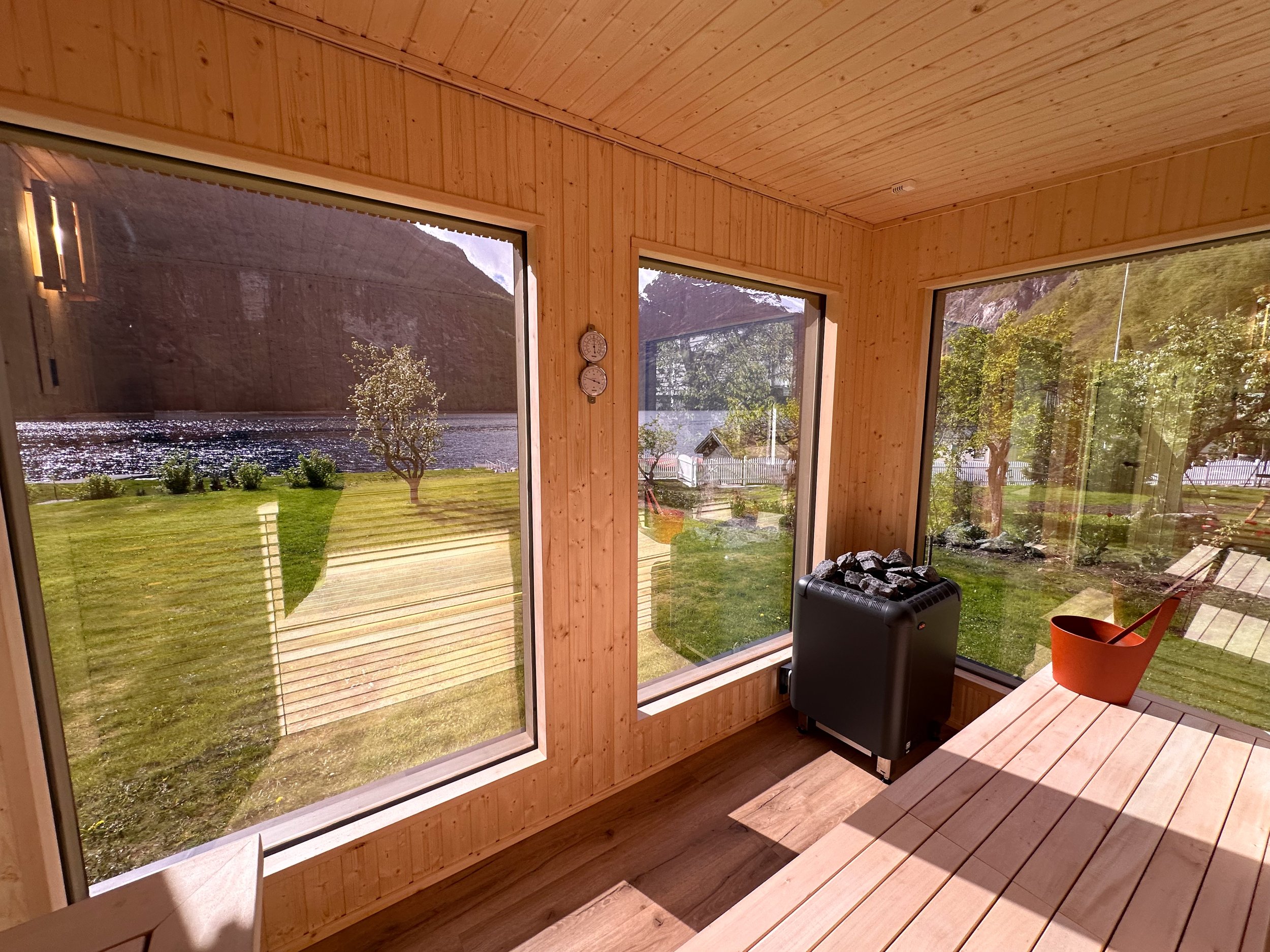 Innendørsområde med store vinduer som viser en utsikt over en hage, fjell, og en innsjø. Det er en privat vedovn eller ovn med kull, og en treskrivebord med en rød gryte på.