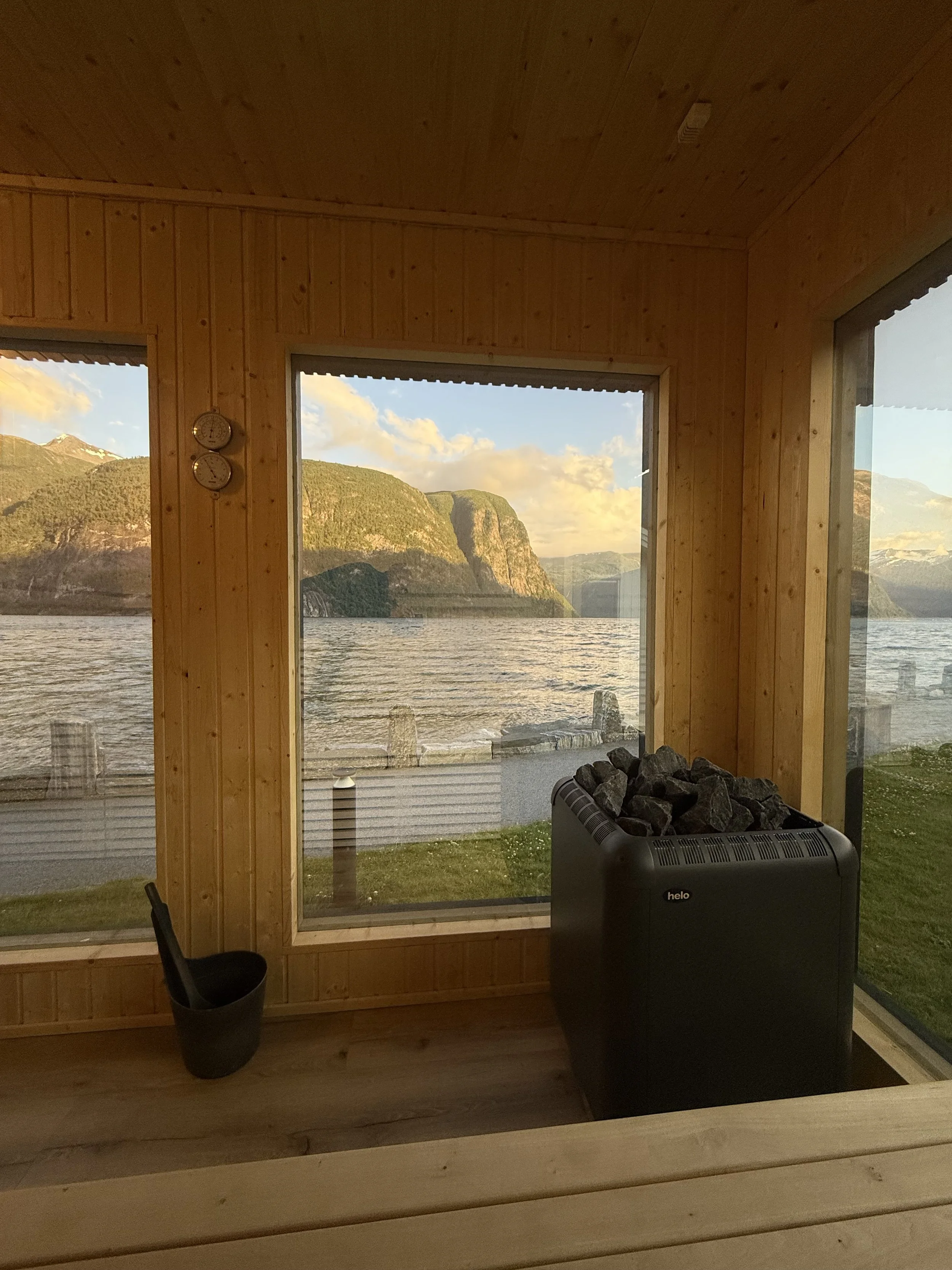 Innendørs wellnessrom med kamin og fjellutsikt gjennom store vinduer, trevegger, og utendørs vann med fjell i bakgrunnen.