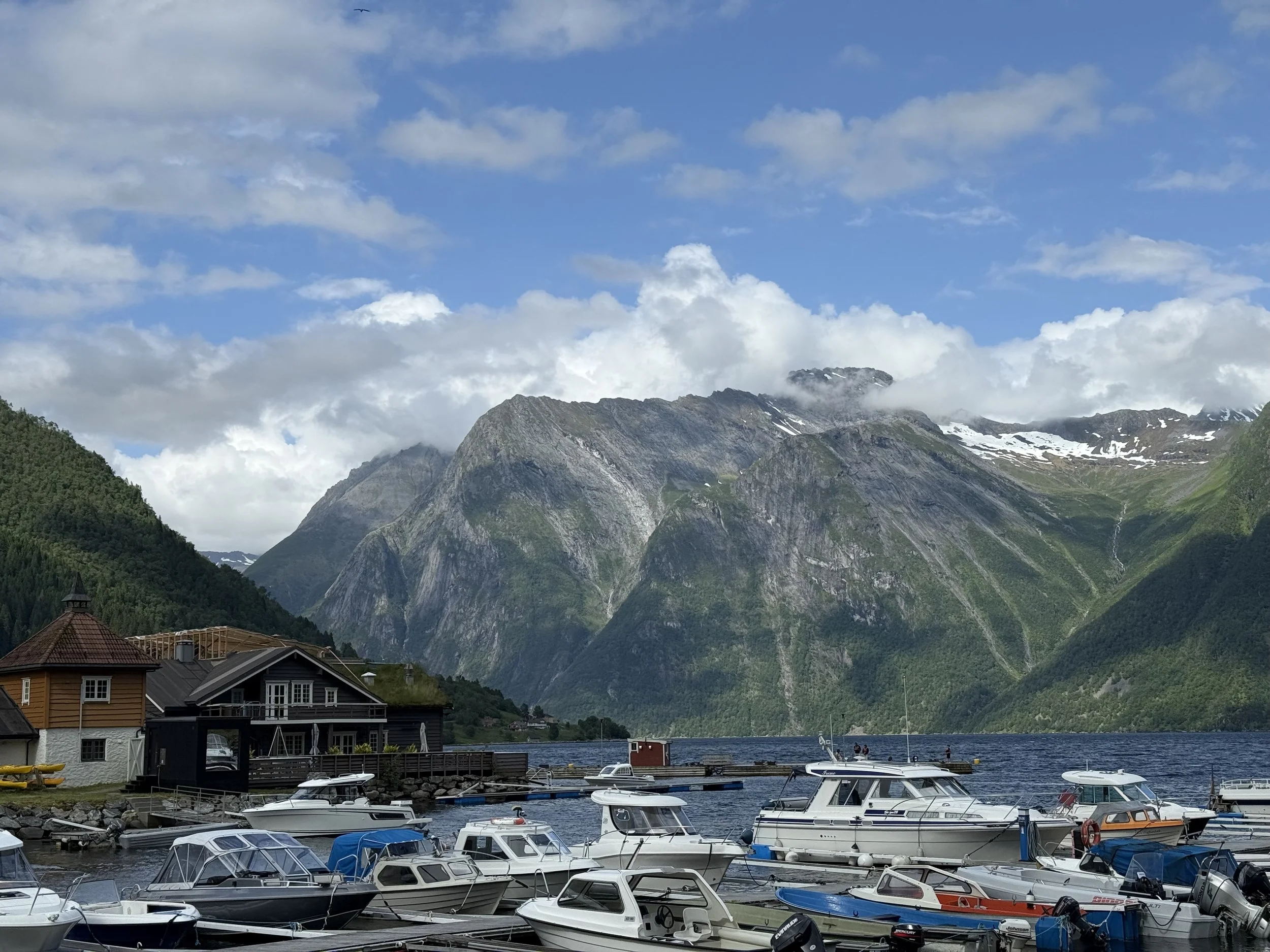 Båter i en havn foran fjell med snø på toppen, omgitt av grønne skråninger og en blå himmel med skyer.