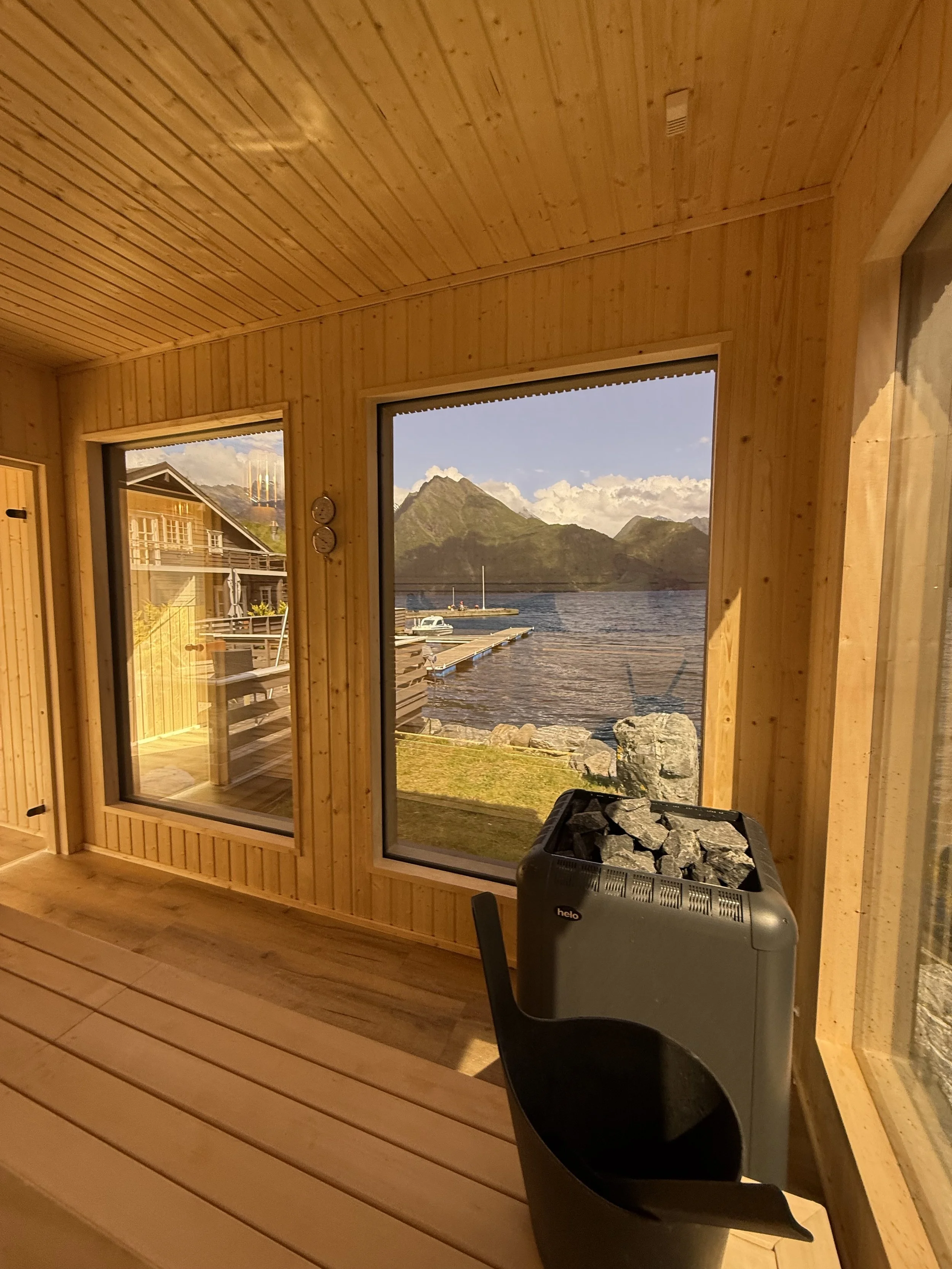 Innendørs med trepaneler, stor vindu med utsikt over fjell og vann, en varmestøtte med steiner, og et lite utendørs område med en bygning i bakgrunnen.