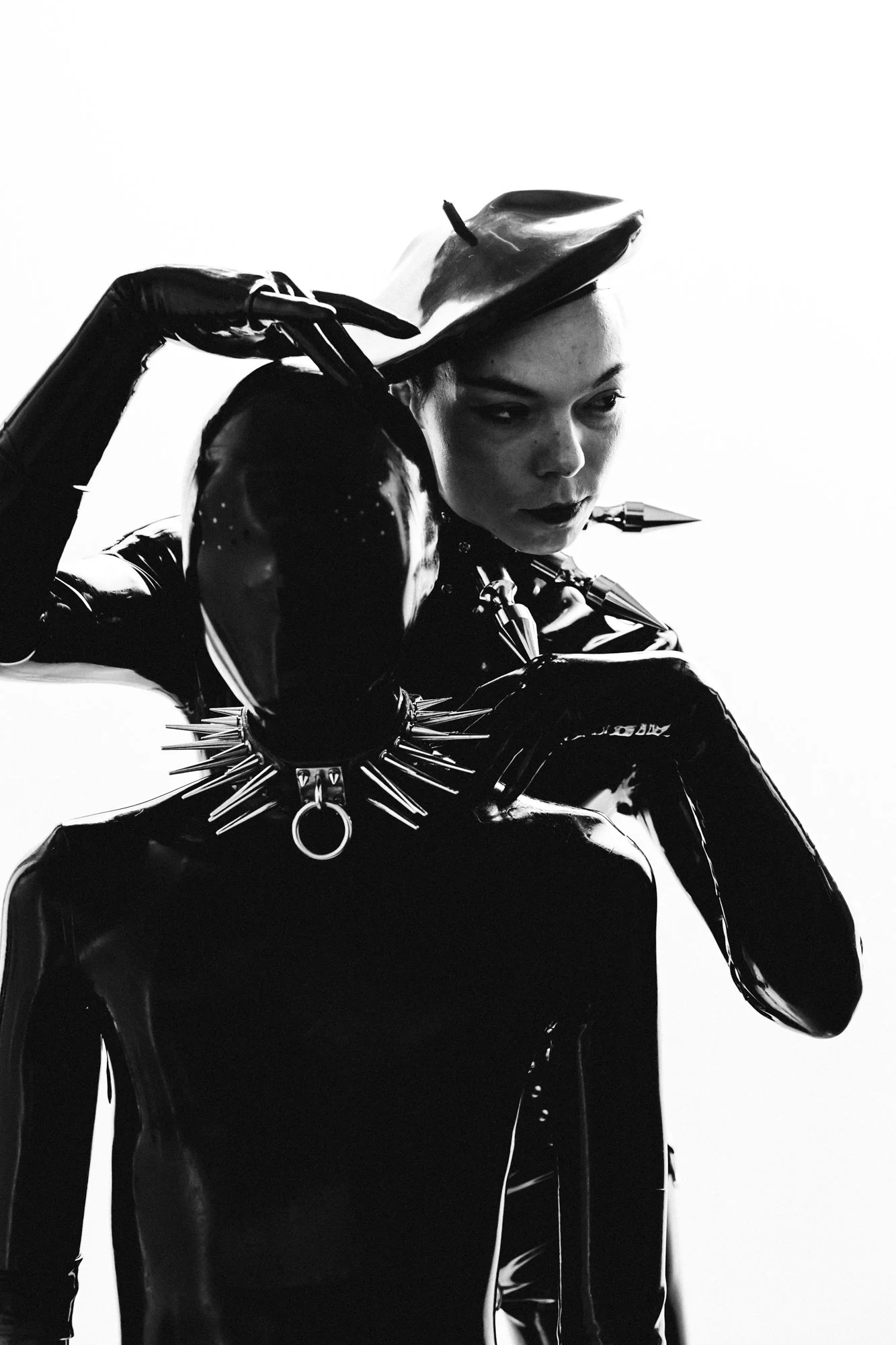 Black and white photograph of a woman with short hair and dark makeup, wearing a shiny black latex outfit with spikes, holding a hat over her head.