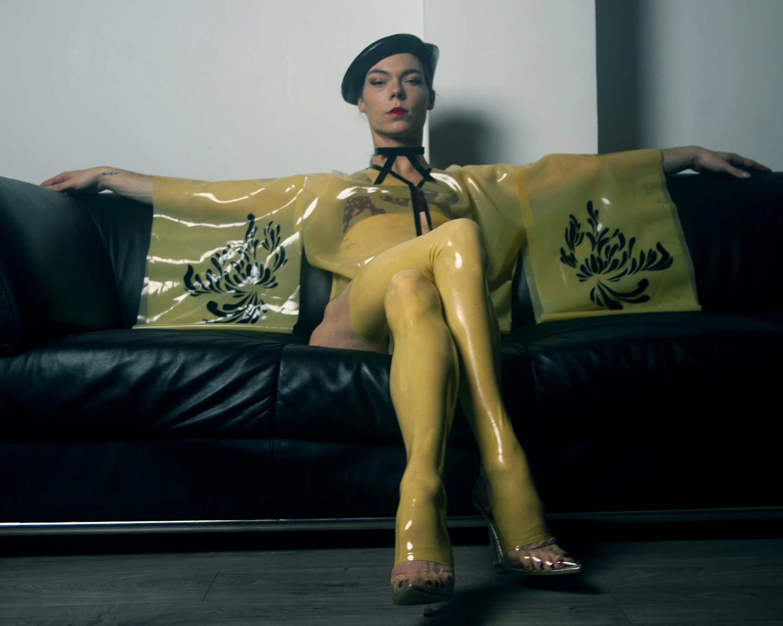 A person sitting on a black leather couch with arms outstretched, wearing yellow latex pants, a yellow blouse with black floral patterns, and a black beret, in a minimalistic room with white walls.