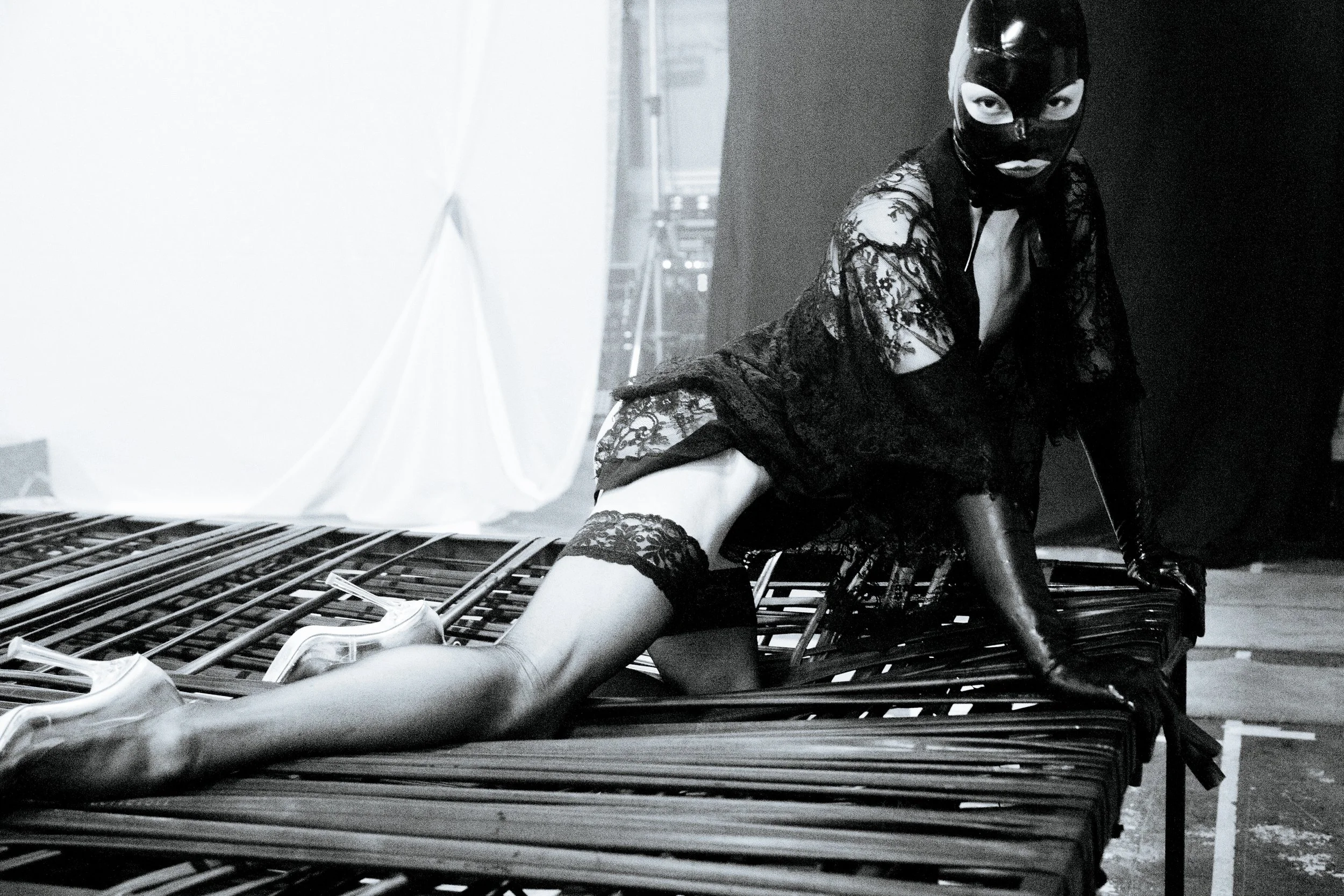 A person wearing a black leather mask and lace clothing sitting on a metal structure in a dark industrial setting.