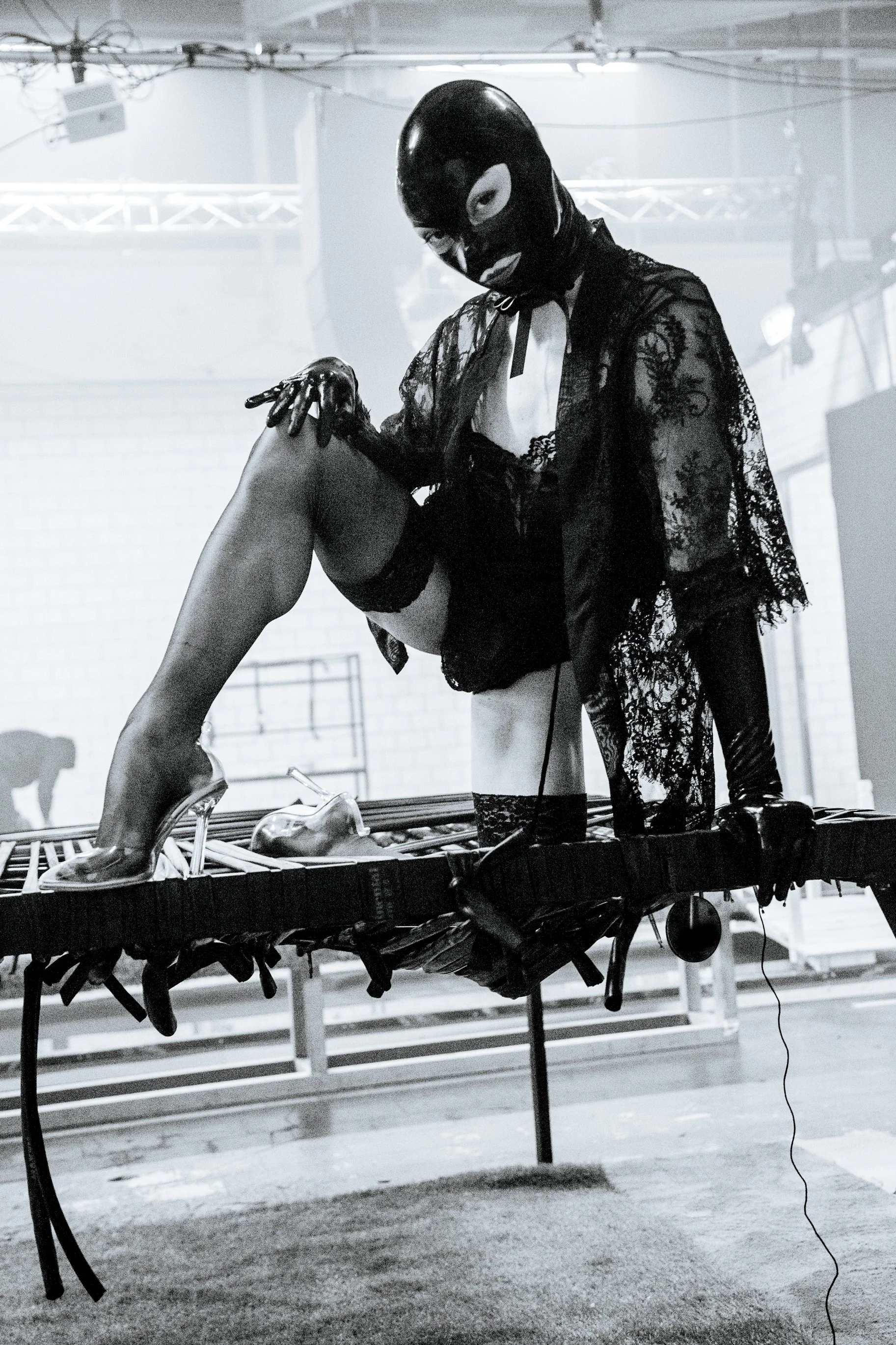 A person wearing a black mask and lace robe stands on a trampoline, holding a woman's leg. The scene appears to be in a gym or studio with equipment and lighting overhead.