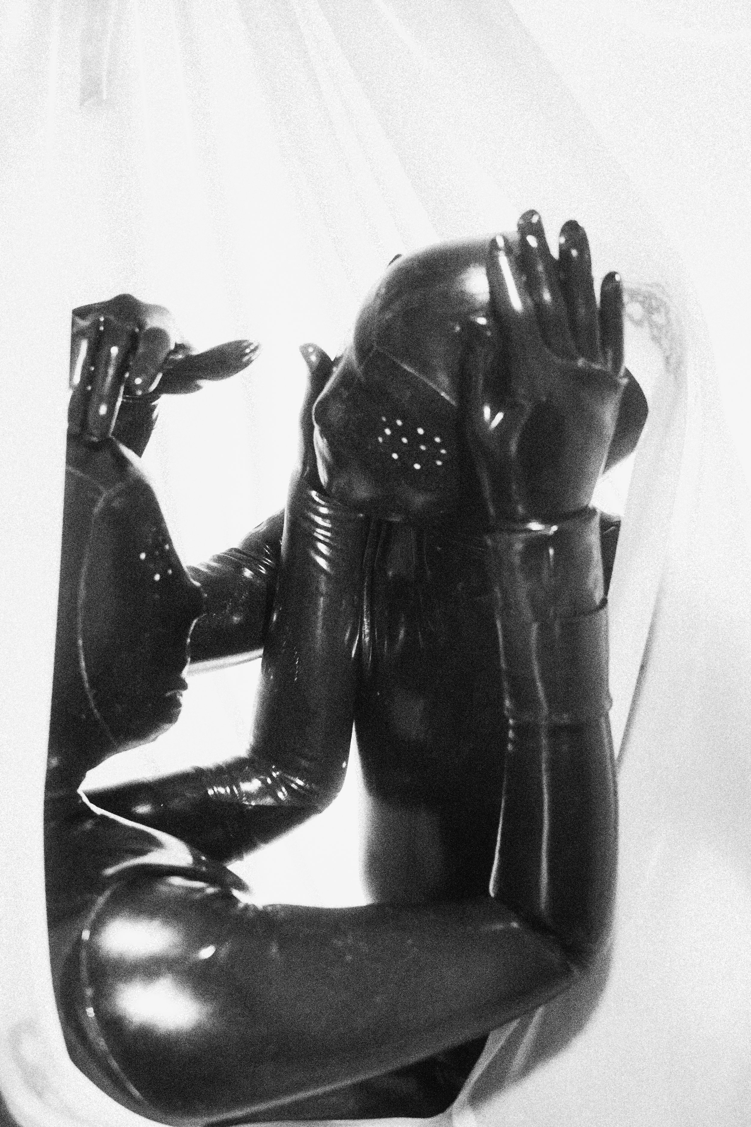 A person wearing a shiny, black leather jacket, gloves, and a helmet, sitting in a car with the window rolled down, holding a smoking cigarette, and making a peace sign with their fingers.