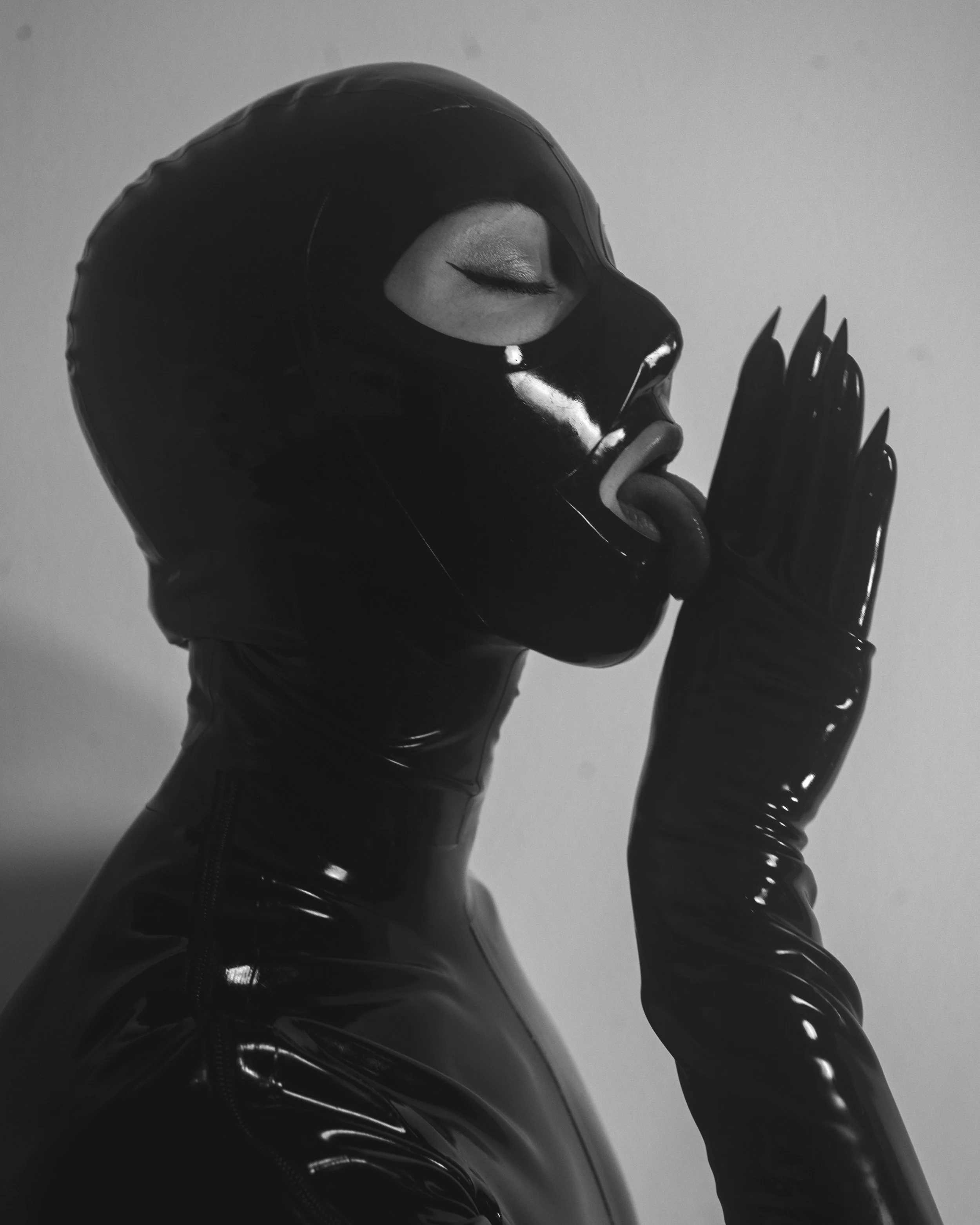 A person wearing a shiny black latex mask with a zipper, black latex gloves, and black latex clothing, with eyes closed and tongue out, in a monochrome setting.