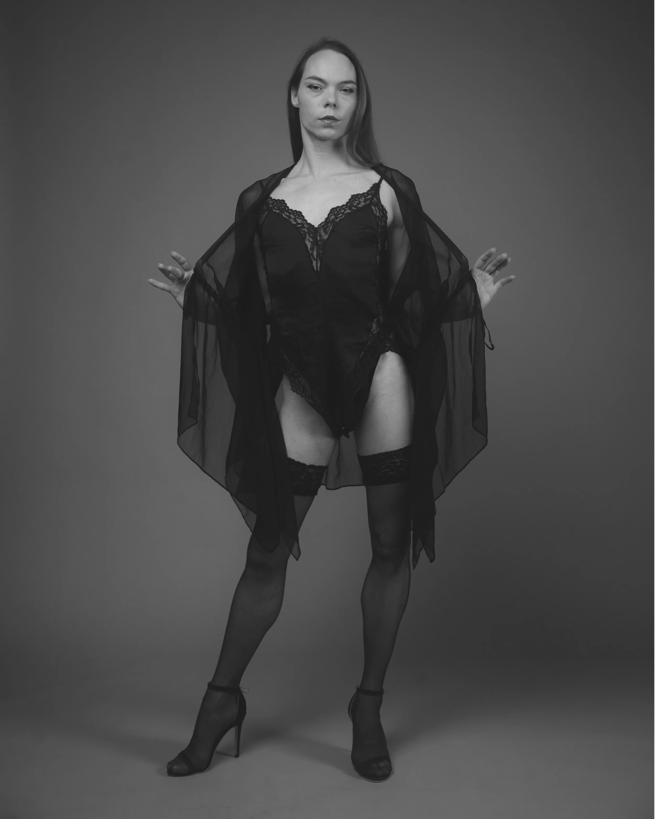 A woman in black lace lingerie and a sheer black robe poses with her arms slightly raised, wearing high heels, against a plain gray background.