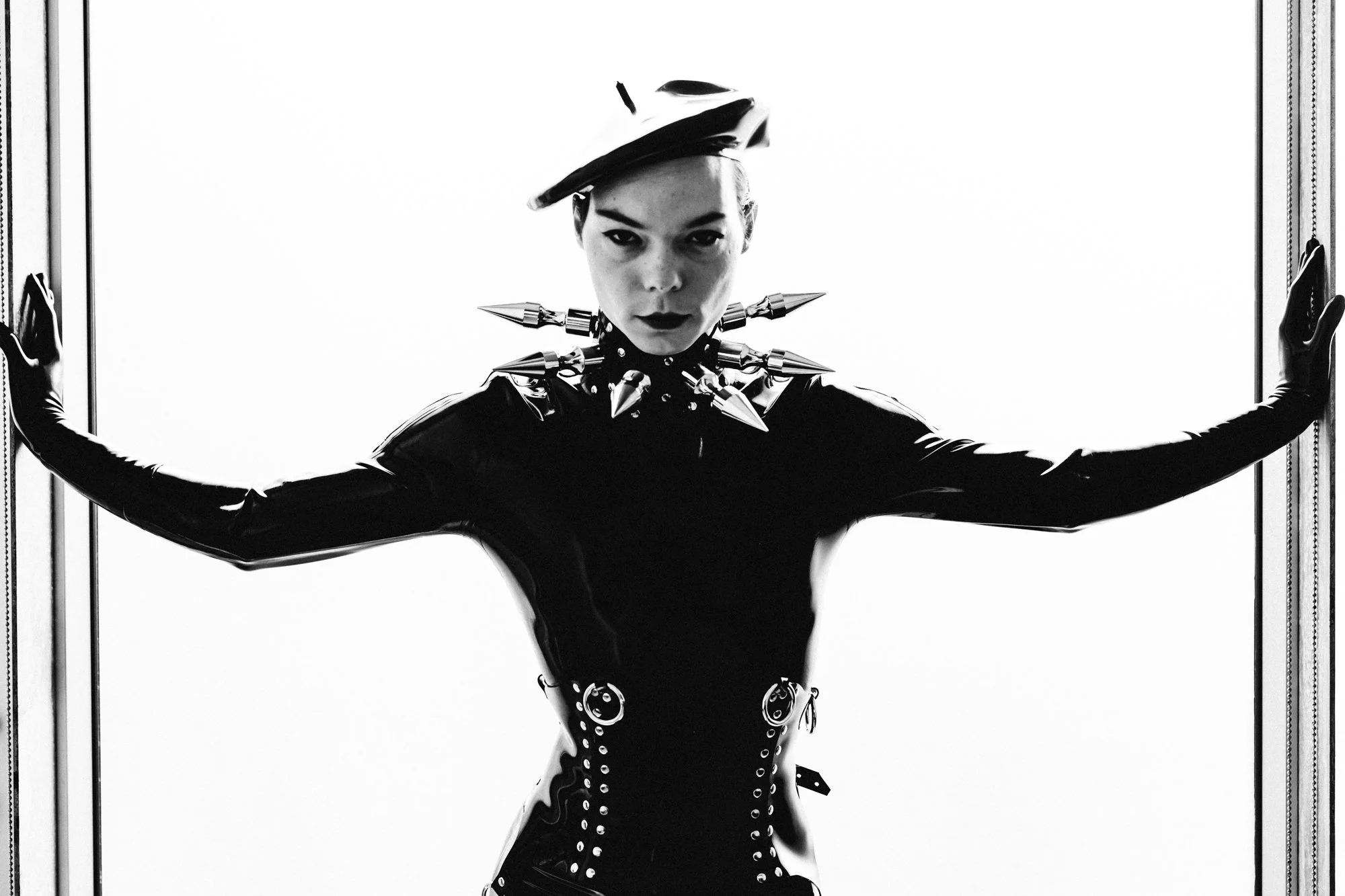 A woman dressed in black latex with spiked metal accessories, wearing a hat, standing with her arms outstretched in front of a white background.