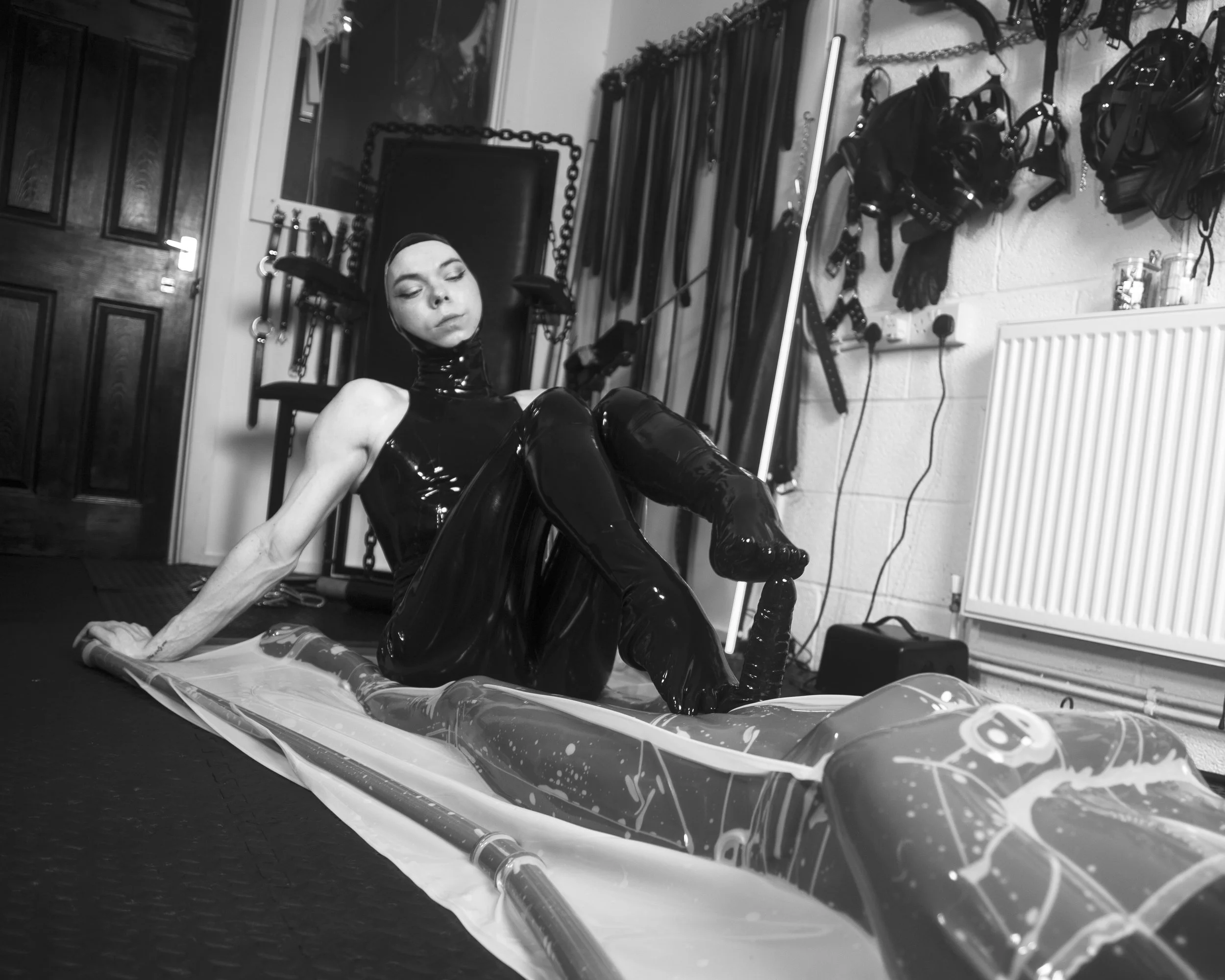 A person dressed in black latex clothing sitting on a skateboard in a room with BDSM gear hanging on the wall.
