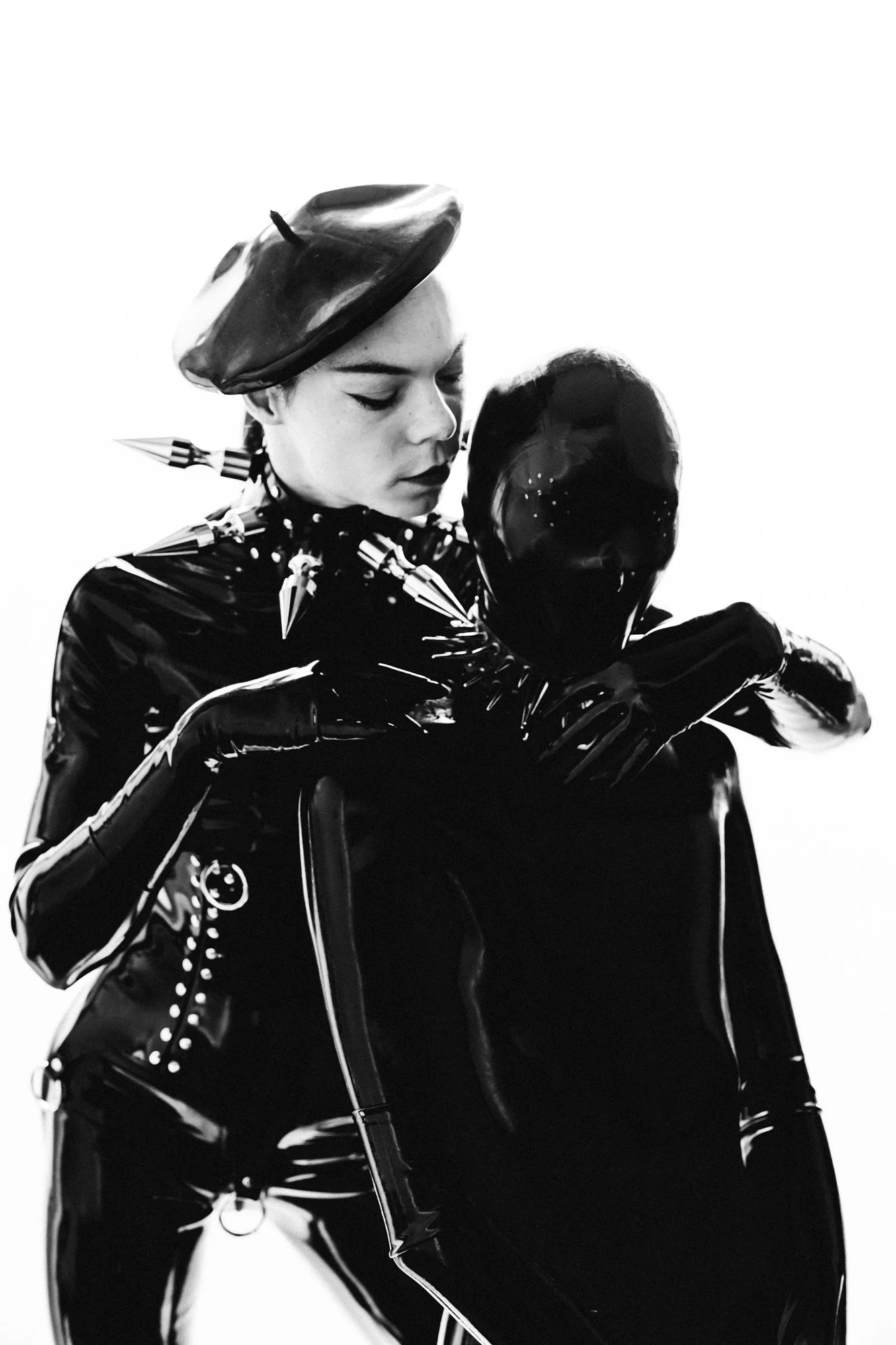A woman dressed in a shiny, latex black suit with spikes on her shoulders and gloves, wearing a beret, kissing a black shiny dog.