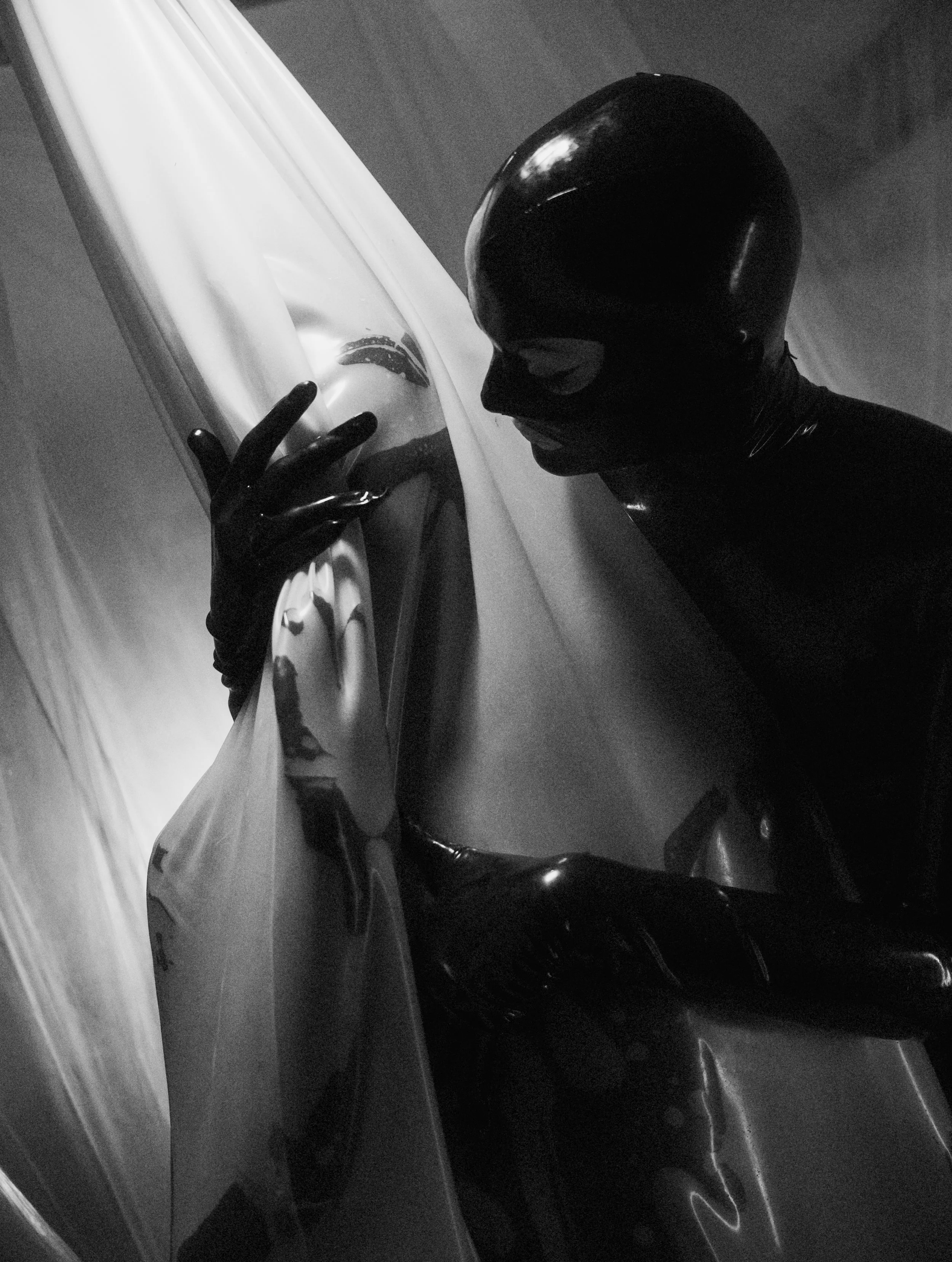 A person dressed in a shiny black latex bodysuit and gloves, wearing a black latex hood with cutouts for the eyes and mouth, poses closely to a reflective metallic surface or sculpture, creating a dramatic, artistic scene in black and white.
